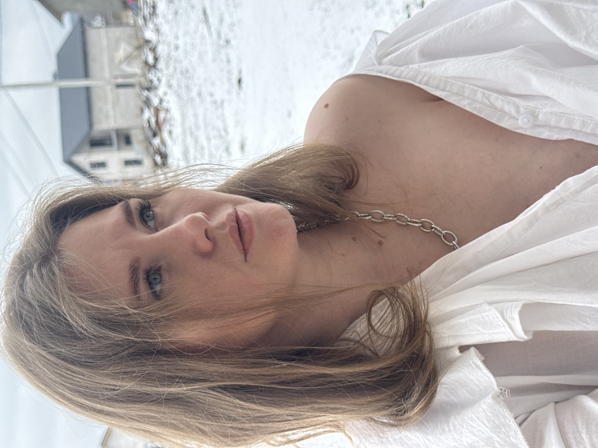 A fair-skinned woman with light brown hair and blue eyes is lying down, facing the camera at an angle. She wears a silver chain necklace and a white shirt that is partially unbuttoned. The background shows a snow-covered ground and a multi-story house. This location is Gornyi (Dvurechenskoe S/P), Russia.