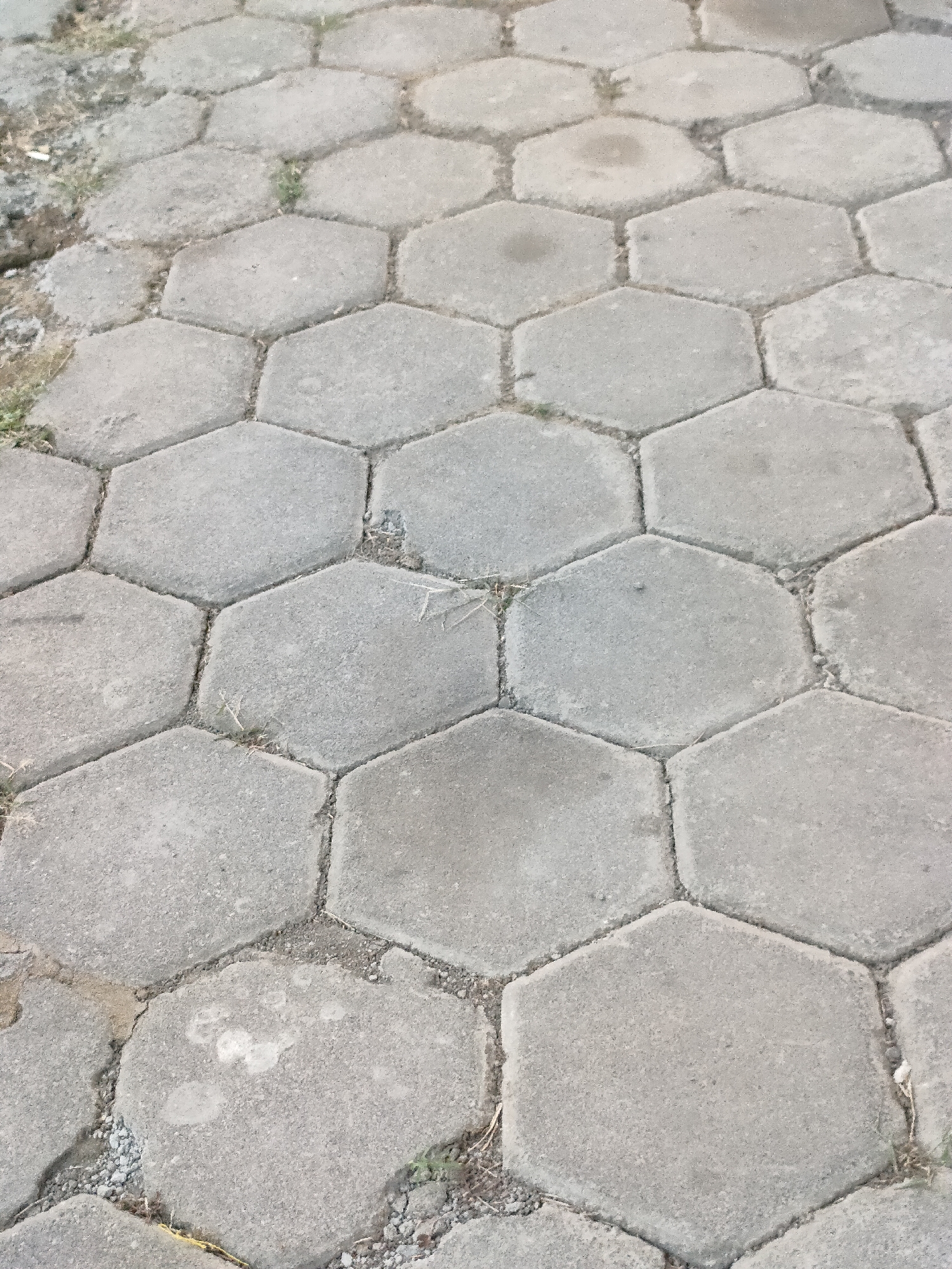 The image displays a close-up, overhead view of a paved ground surface composed of interlocking hexagonal concrete pavers. The pavers are a light gray color, with some exhibiting slight variations in tone and texture, suggesting weathering and age. The spaces between the pavers are filled with a mixture of dirt, small pebbles, and sparse blades of dry grass, indicating an outdoor environment. The paving is arranged in a repeating geometric pattern that recedes into the background, suggesting a pathway or courtyard. No people, animals, or vehicles are visible in the frame. The lighting appears to be natural daylight, casting subtle shadows that highlight the texture of the pavers. The scene is situated in Banguntapan, Indonesia, and depicts a common urban or suburban ground surface material.