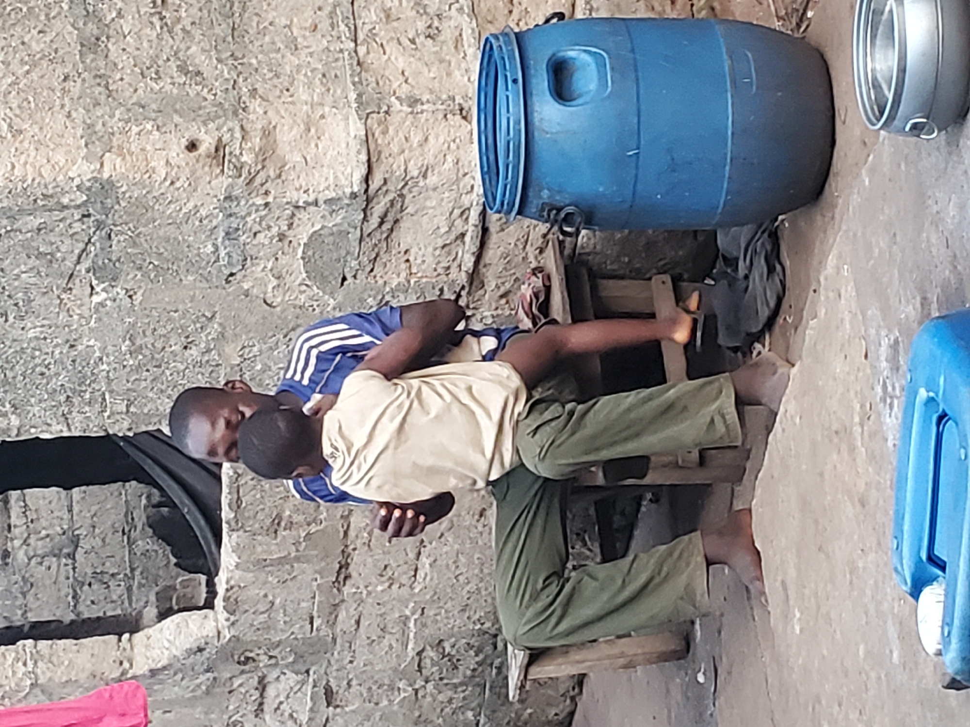 The image shows two individuals in an outdoor setting in Ukwu-Abwa, Nigeria. One individual, an adult male wearing a blue shirt with white stripes, is bent over and appears to be attending to a child. The child is wearing a light-colored shirt and green pants, and is sitting on a makeshift wooden structure. Next to the structure is a large blue plastic barrel. In the background is a rough stone wall, and on the ground are various other objects including a metal container and a blue plastic container.