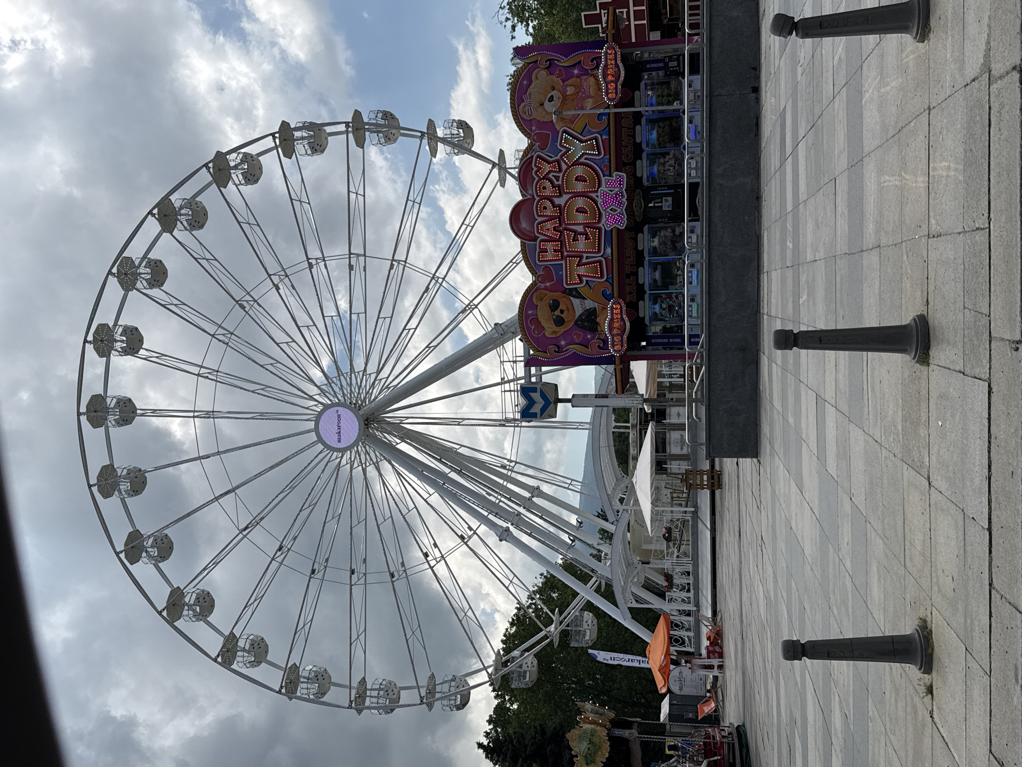 The original image, which appears to be captured in portrait orientation but is displayed rotated on its side, depicts an outdoor amusement area in Sofia, Bulgaria. The central subject is a large, white Ferris wheel, its intricate metal structure and numerous passenger gondolas dominating the left side of the frame. The hub of the Ferris wheel prominently displays the text "makaroon".
To the right of the Ferris wheel is a vibrant game booth named "HAPPY TEDDY", illuminated with lights and adorned with graphics of teddy bears. Visible text on the booth includes "BIG PRIZES", "WINNING", "XXL TEDDY", and "BOSS" (associated with a teddy bear wearing sunglasses). Display cases below the main sign show various plush toys and prizes.
The ground is paved with light grey rectangular blocks, and several dark grey, cast-iron bollards are placed along the path. In the background, patches of green trees are visible beneath a partly cloudy sky with hints of blue. Further back on the right, white outdoor umbrellas, tables, and chairs suggest a cafe or restaurant, possibly named "BELA VISTA" as indicated by OCR. An orange flag or banner also bears the text "makaroon.bg". A blue "M" symbol, characteristic of a metro station sign in Sofia, can be seen near the base of the Ferris wheel, providing a strong location cue.
The scene suggests a lively atmosphere typical of a fairground or public entertainment space, likely captured during the daytime on a bright, partly cloudy day. While no people are actively visible, the setup implies a recreational event or leisure activity.