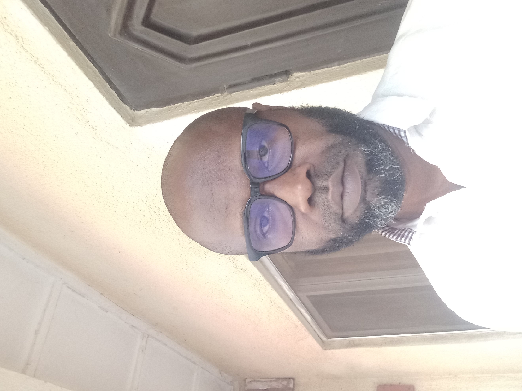 A bald, bearded man with dark skin and glasses is facing the camera. He is wearing a white shirt. The background includes a ceiling with a dark brown inset panel and a textured wall. The location is Barden Yana, Nigeria.