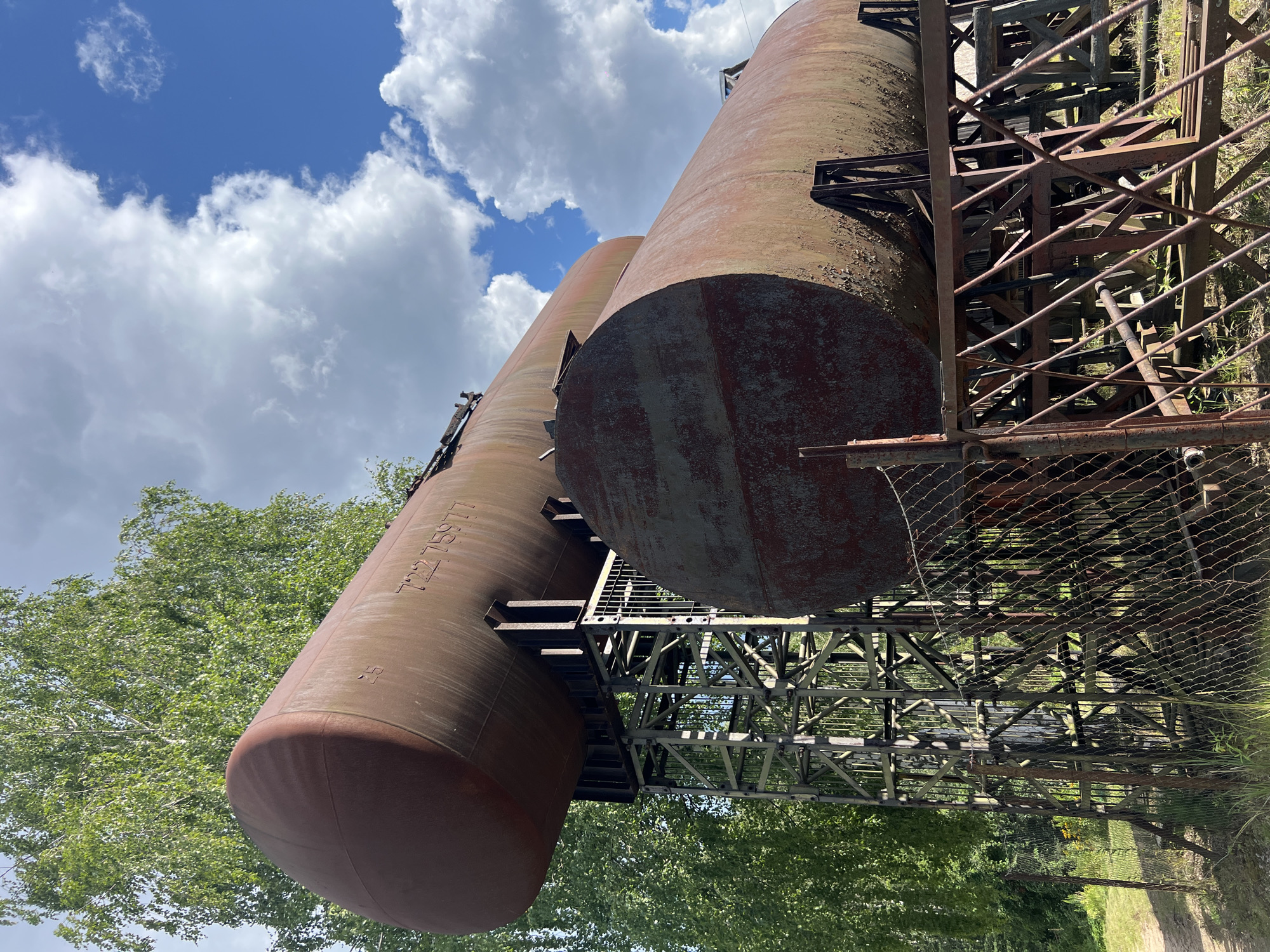 Two large, rusty, cylindrical storage tanks are supported by a metal structure in an outdoor environment. The tanks are positioned one above the other, with the upper tank appearing larger and longer than the lower tank. Both tanks show signs of significant corrosion and wear. The supporting structure is made of welded metal beams and crossbars, forming a robust framework. In the foreground, a chain-link fence runs across the scene, partially obscuring the view of the tanks and their supports. Beyond the fence, there is overgrown grass and vegetation, suggesting an unkempt or abandoned area. In the background, a dense green tree with light-colored bark stands to the left, and a blue sky with scattered white clouds is visible overhead. The scene appears to be located in a rural or industrial area. The location is Strakhovo (Sitnikovskii S/S), Russia. There are no people visible in the image, and no actions or interactions are taking place.