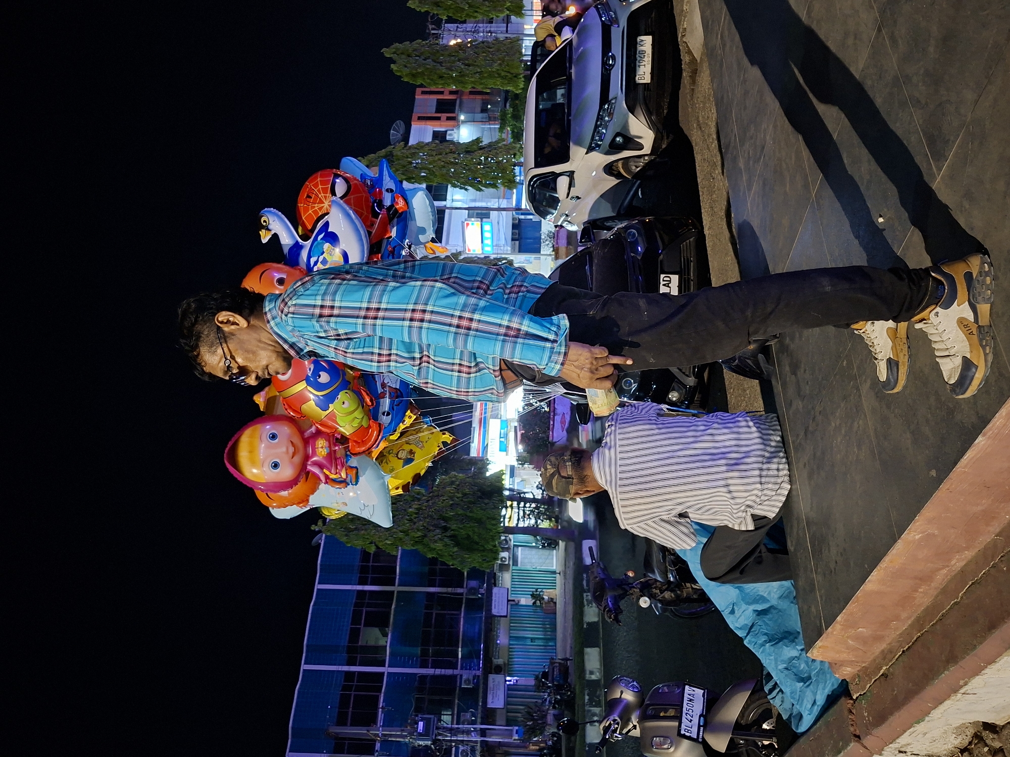 The image depicts two men interacting in a brightly lit urban environment. One man, wearing a blue plaid shirt and black pants, holds a large assortment of cartoon character balloons. He appears to be selling them. The other man, wearing a striped shirt and what appears to be turquoise pants, is counting out money. The scene occurs on a sidewalk near a street, where several motorcycles and a silver car are parked. The building in the background and the presence of vehicle registration plates indicate the location is Lhokseumawe, Indonesia. The environment suggests a nighttime setting with businesses or shops nearby.