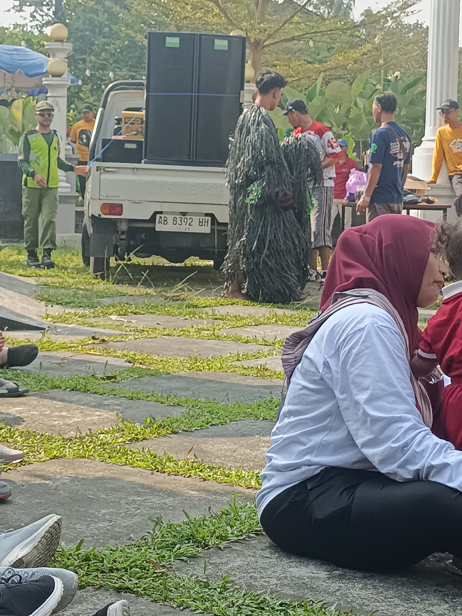 A group of people are gathered outdoors in an urban environment, likely in Sleman, Indonesia, possibly for an event or performance. In the foreground, a person wearing a maroon hijab and a white long-sleeved shirt sits on the ground, facing right, with black pants. To their right, another person in a red outfit is partially visible. Behind them, a person in a dark, leafy costume stands and interacts with another individual in a white and red shirt.
A white Mitsubishi pickup truck is parked, loaded with large black speakers. A man in a yellow shirt and a green vest stands next to the truck, holding a blue rope. Another man in a yellow t-shirt stands further back. The ground is a patterned walkway with sections of grass. In the background, trees and foliage are visible, along with a white pillar and part of a blue awning. The lighting suggests it is daytime, with sunlight casting shadows.