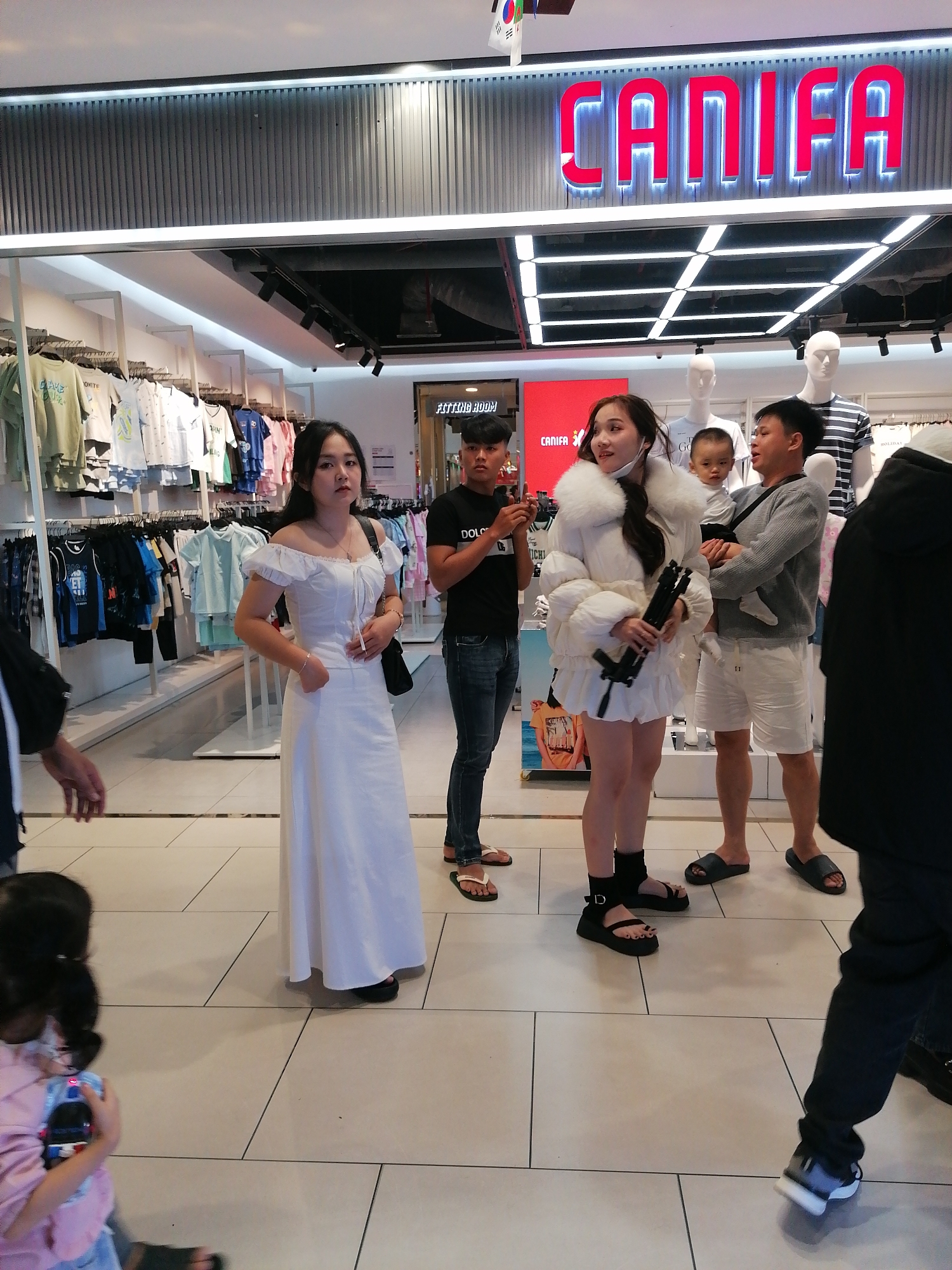 The image shows a group of people inside a clothing store. In the foreground, a young woman in a white dress stands facing the camera, with a black purse slung over her shoulder. To her right, another young woman in a white puffer jacket and a short skirt is holding a black object that resembles a selfie stick or a small tripod. Behind them, a man is holding a baby, and a young boy is also visible. Further in the background, clothing racks are filled with various garments. The store appears well-lit with bright overhead lighting. A prominent sign with red illuminated letters reads "CANIFA". The setting is indoors, likely a commercial establishment, given the presence of merchandise and mannequins. The overall atmosphere suggests a casual shopping environment. The time of day is not discernible, and there are no indications of weather. The location cue "Da Lat, Vietnam" provides geographical context.