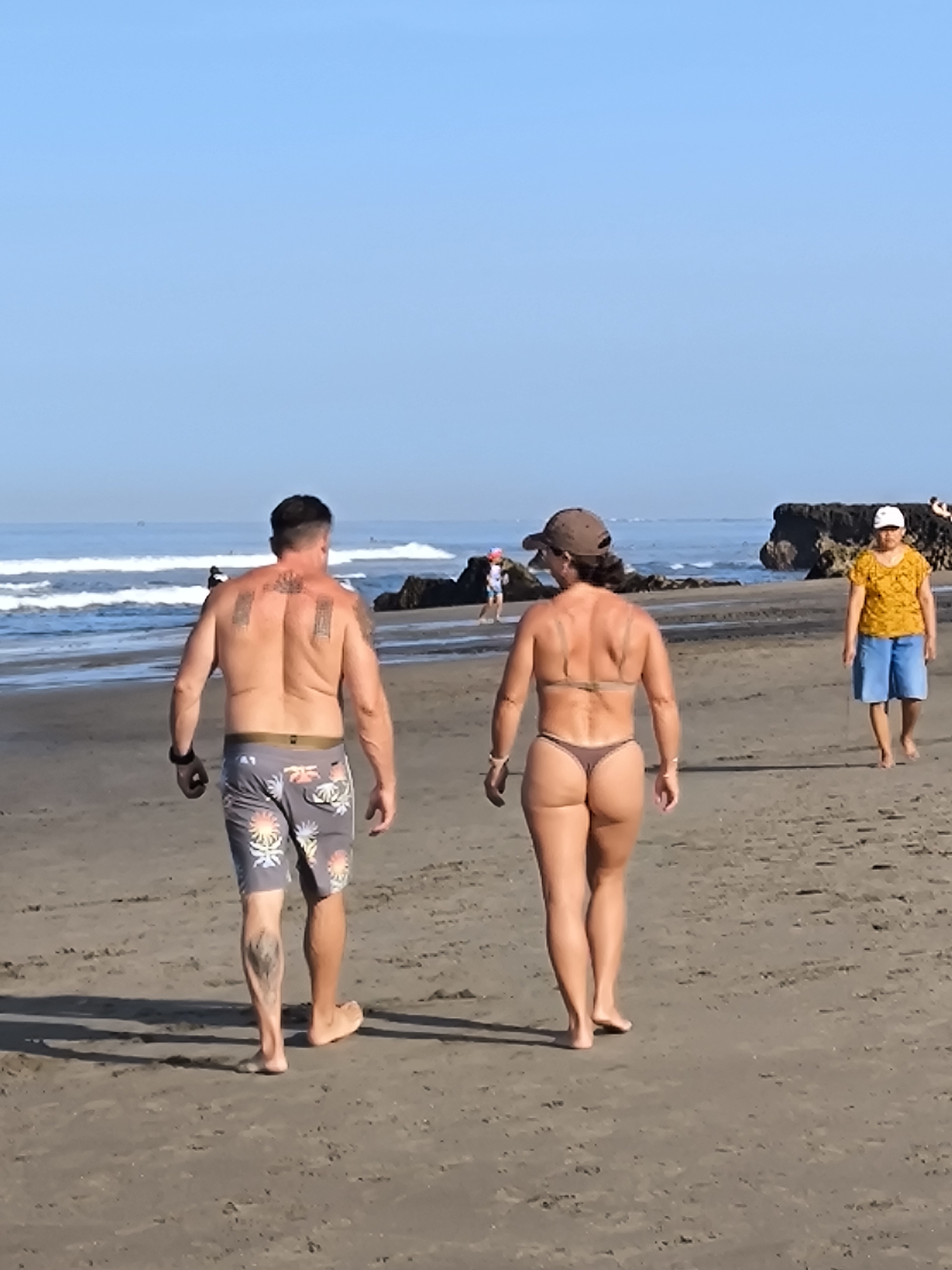 The image captures a scene on a beach in Canggu, Indonesia. Two individuals, a man and a woman, are walking away from the camera. The man is bare-chested with tattoos on his back and legs, wearing patterned shorts and a black wristwatch. The woman, walking next to him, is wearing a bikini and a baseball cap. A few other people are visible in the background, including a person in a yellow shirt and blue shorts walking on the beach. The ocean with visible waves is on the left side, and dark rock formations are present further down the beach. The sky is clear and blue.