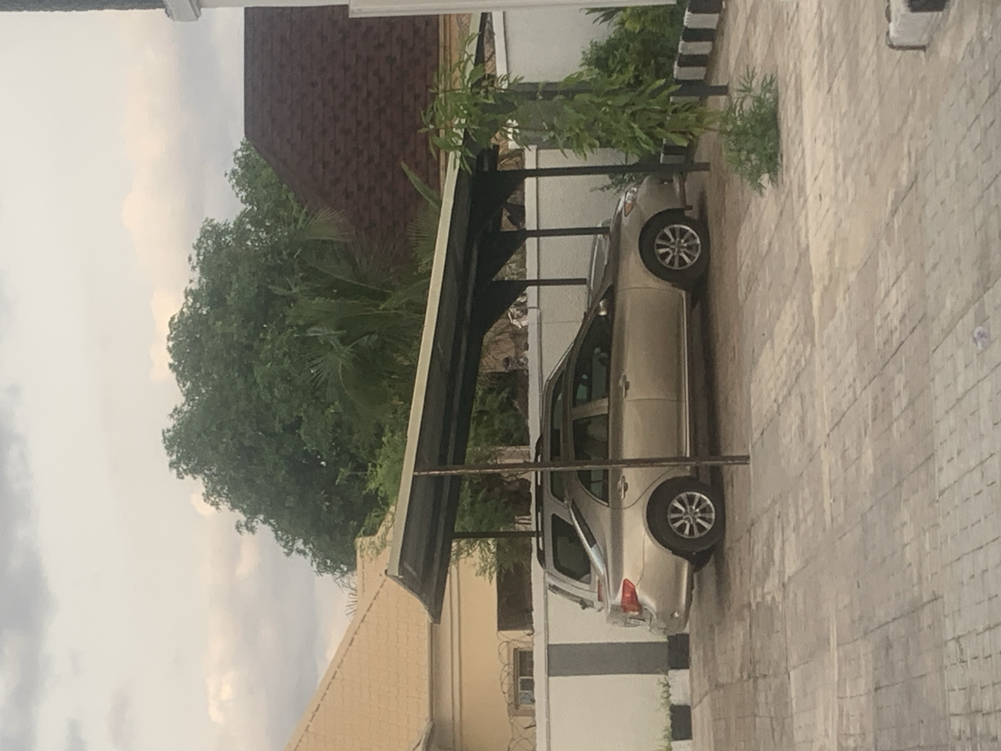 A gold sedan is parked under a grey carport in a paved lot. The sedan is facing left and has its front and rear wheels visible. Behind the sedan, another vehicle, possibly an SUV, is partially visible. The carport has a curved, dark grey roof supported by thin, dark grey poles. The area around the carport is paved with light brown rectangular stones. To the left of the carport, a low white wall with a barbed wire fence on top separates the lot from a building with a pale yellow, tiled roof. To the right of the carport, a thin, tall green plant grows near a white wall with black and white striped borders. In the background, dense green trees and the dark red, tiled roof of another building are visible under a cloudy sky. The scene appears to be in a residential or commercial area of Jalingo, Nigeria. There are no people visible in the image.