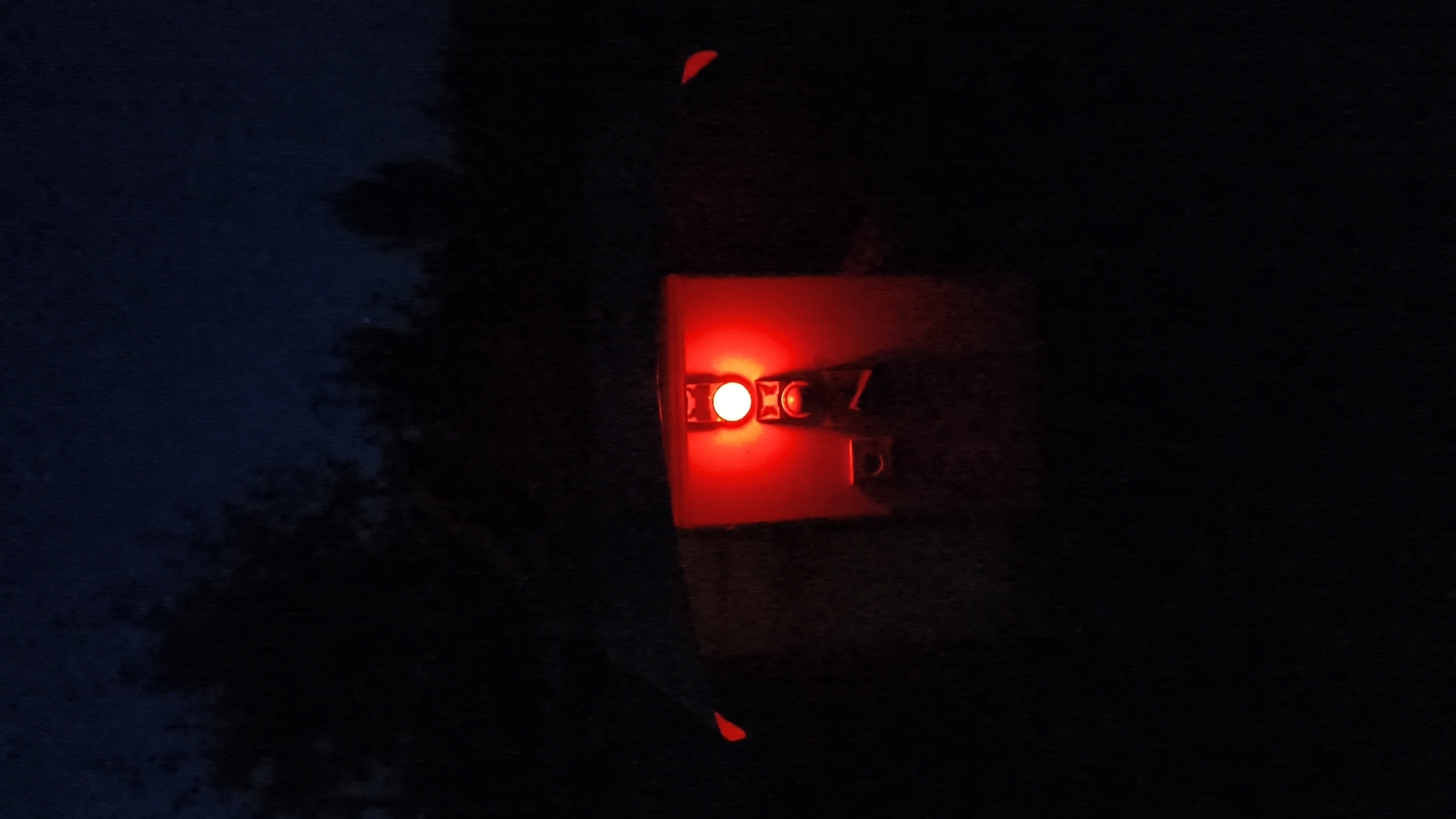 The image, taken at night in an outdoor, unlit environment, shows a close-up view of a red object with a bright, glowing red light at its center. The object appears to be a safety light or reflector, likely attached to a bicycle or other equipment, positioned low to the ground. The light is circular and emits a strong, diffused glow, illuminating the immediate surroundings. To the left and right of the central light, there are smaller, also red, wing-like protrusions. The object is mounted on a darker, rectangular base. The background is dark and indistinct, with silhouettes of trees or foliage visible against a deep blue night sky. There are no people or animals in the frame.