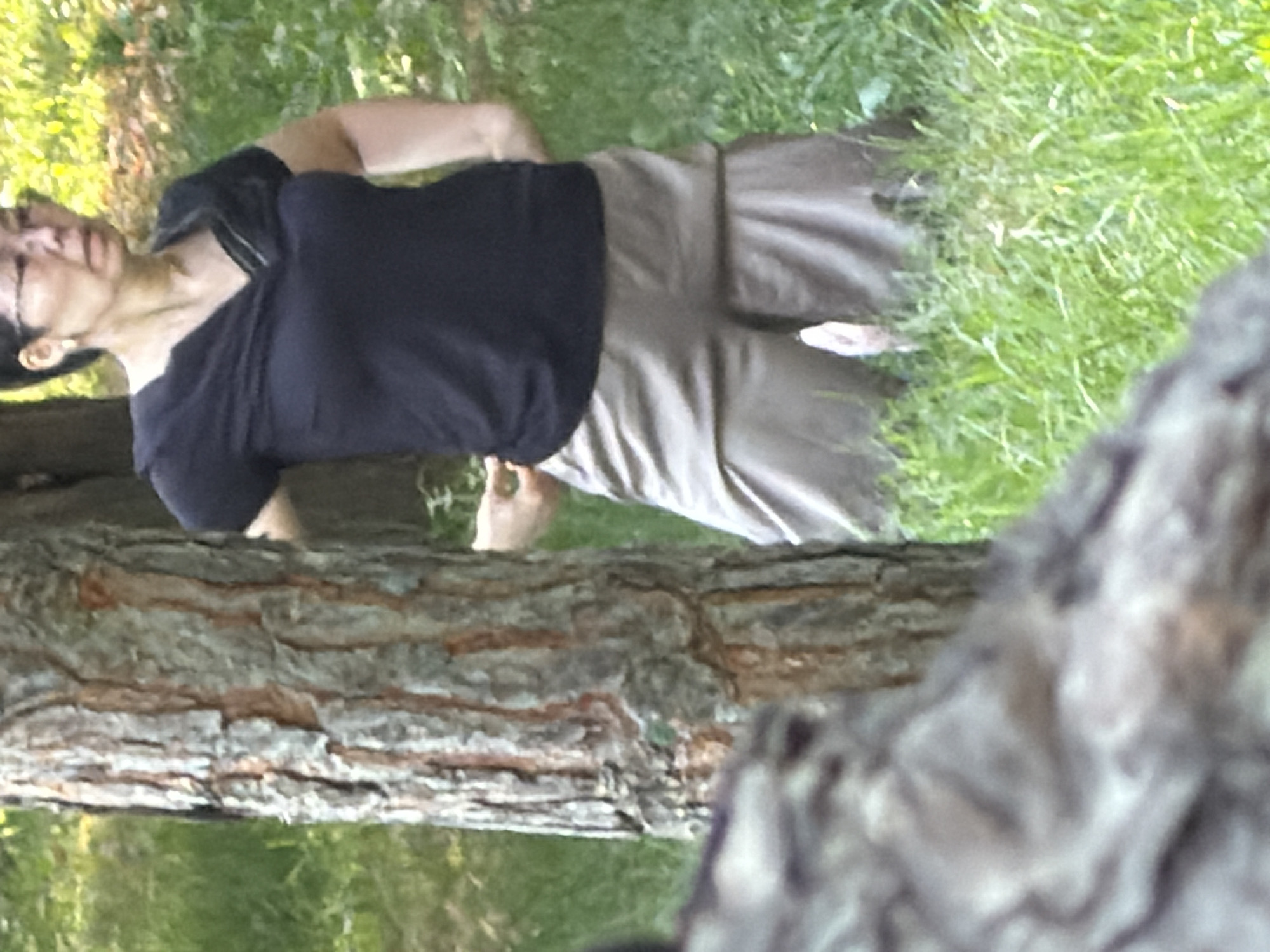 A woman stands in front of a tree, wearing a black shirt and beige pants. She is wearing glasses and appears to be looking to her right. The background consists of grass and foliage, suggesting an outdoor environment. The tree trunk is visible to the left of the woman, and a blurred foreground element, possibly another tree or log, is on the far left.