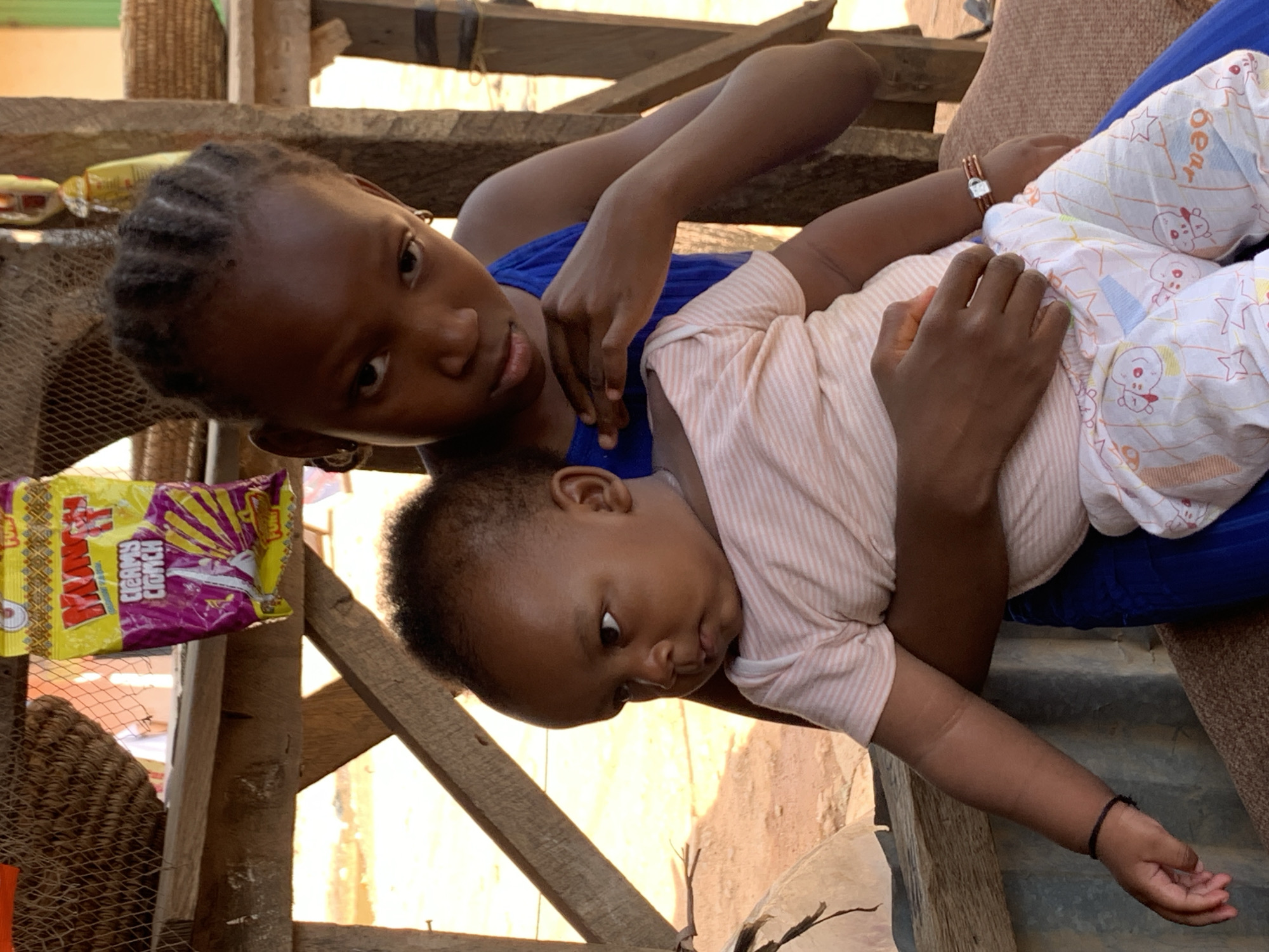 In Sabon Tasha, Nigeria, an older girl with braided hair and a blue top holds a baby wearing a pink striped shirt and patterned pants. They are positioned indoors, perhaps on or near wooden stairs, as evidenced by wooden planks and structures in the background. To the left is a yellow and purple package, possibly a snack item. The setting suggests a humble dwelling. The older girl looks directly at the viewer, while the baby appears to be looking down.