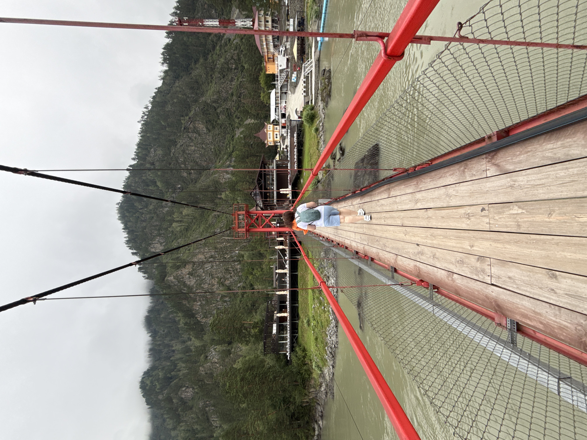 A woman with a backpack walks across a suspension bridge over a river. The bridge has red metal railings and a wooden walkway. In the background, there are green mountains covered with trees, and a cluster of buildings on the riverbank, including a restaurant and houses. The sky is overcast. The location is identified as Barangol, Russia.