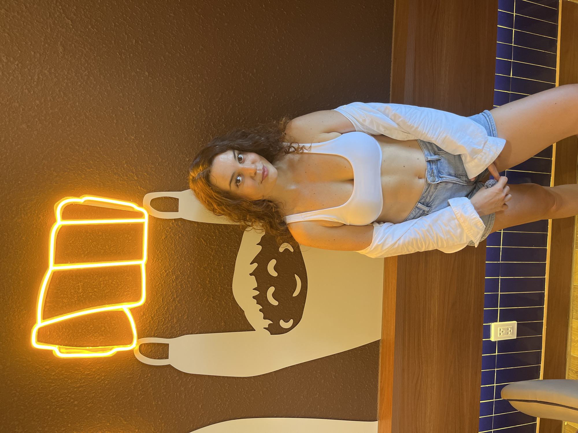 A woman stands in front of a wall with a brown textured finish. A neon light fixture in the shape of a roll of film is illuminated in yellow above a graphic of a hand holding a smiling cartoon face. The woman is wearing a white crop top, denim shorts, and an unbuttoned white shirt that is draped off her shoulders. She is looking at the camera with a slight smile. The lower portion of the image shows a wooden shelf with blue tiled panels underneath. An electrical outlet is visible on the tiled wall. The scene appears to be indoors, possibly in a restaurant or cafe, with the mural suggesting a connection to filmmaking or photography. The location is identified as Phan Thiết, Vietnam.