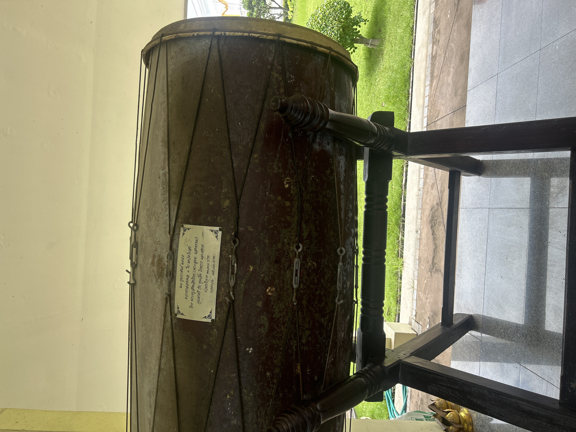 The image shows a large, dark brown drum on a wooden stand, positioned outdoors. A white plaque with Thai text is attached to the drum. The background includes a grassy area with some greenery and a building with a cream-colored wall. The drum is oriented horizontally and appears to be made of wood and metal. The stand is dark wood and has decorative carved elements. The ground beneath the drum and stand is tiled. The setting suggests a public or cultural space.