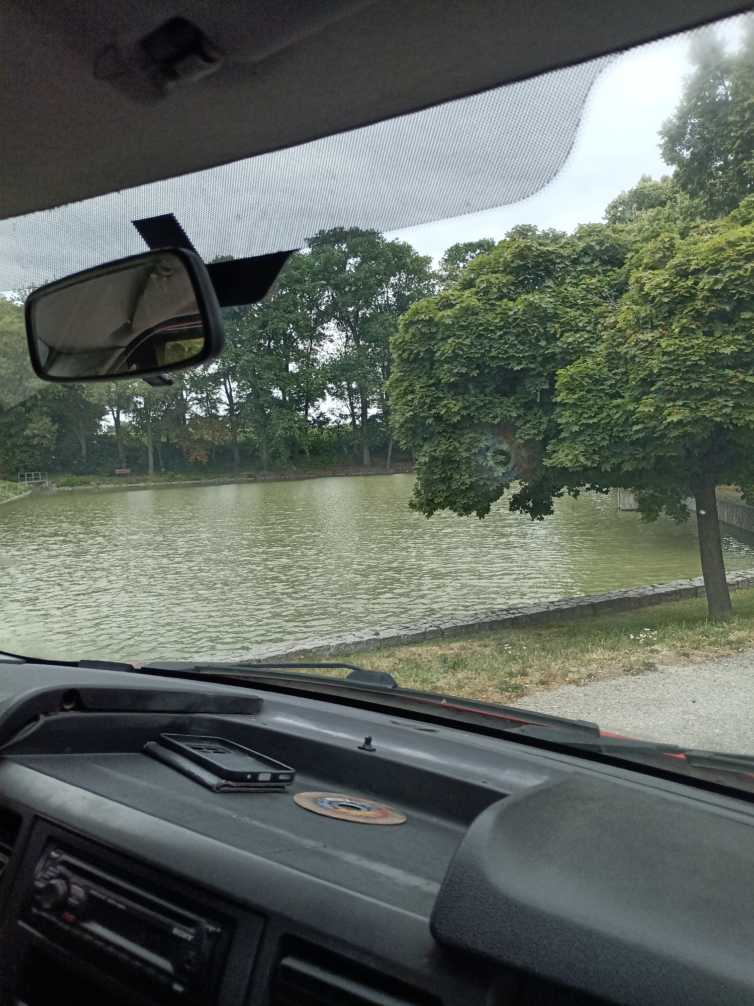 This media file provides a view from inside a vehicle, looking out through the windshield. The scene depicts a calm body of water, likely a pond or small lake, bordered by a stone embankment and lush green trees. In the foreground, the dashboard of the car is visible, with a mobile phone and a small, circular metal object placed on it. A car's rearview mirror is also in view, reflecting the sky and trees.
The weather appears to be overcast, suggesting a cloudy day. The time of day is not explicitly clear but seems to be daytime. The location is identified as Svémyslice, Czechia.
There is no discernible activity or event taking place; the scene is static and peaceful. Notable details include the texture of the water, the dense foliage of the trees, and the arrangement of objects on the dashboard. The overall impression is one of a quiet moment observed from the comfort of a vehicle, possibly during a scenic drive or a stop to enjoy the view. No visible text is present in the image.