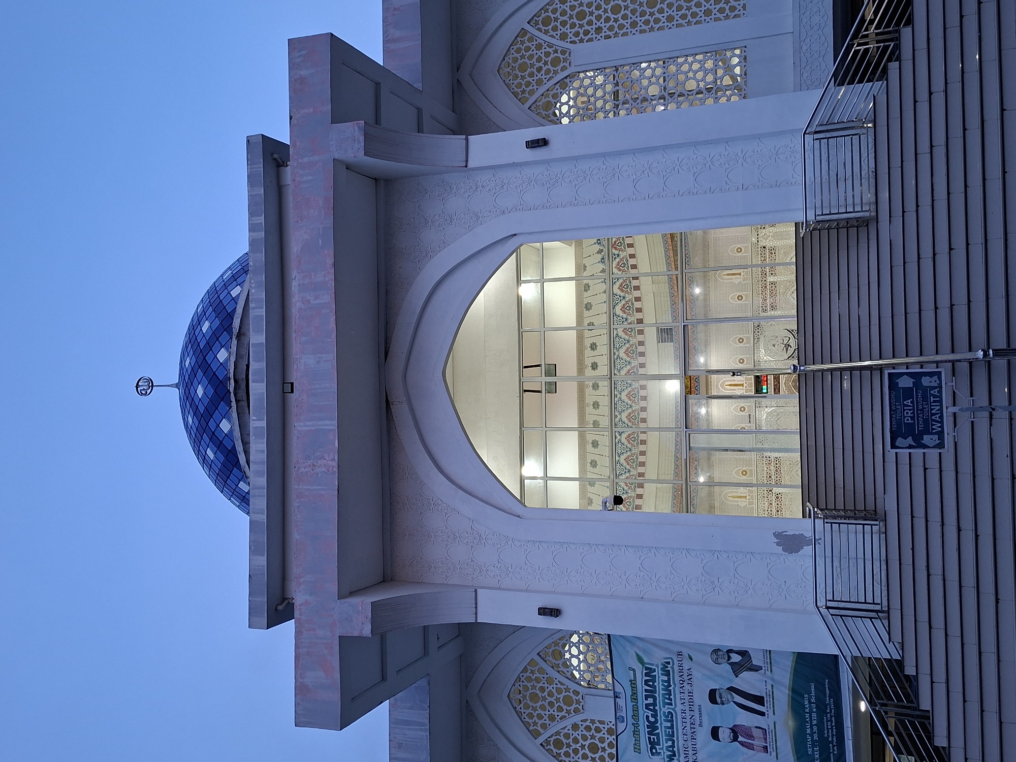 The image shows the exterior of a building, likely a mosque, located in Paya Tringgadeng, Indonesia. The building has a blue and white domed roof, a white arched entrance with intricate patterns, and glass doors leading inside. To the right of the entrance are stairs with metal railings, and a sign indicating separate ablution facilities for men (PRIA) and women (WANITA). A banner advertising a "PENGAJIAN MAJELIS TAKLIM" (Islamic Studies Assembly) is visible at the base of the stairs. The sky is a clear blue in the background.
