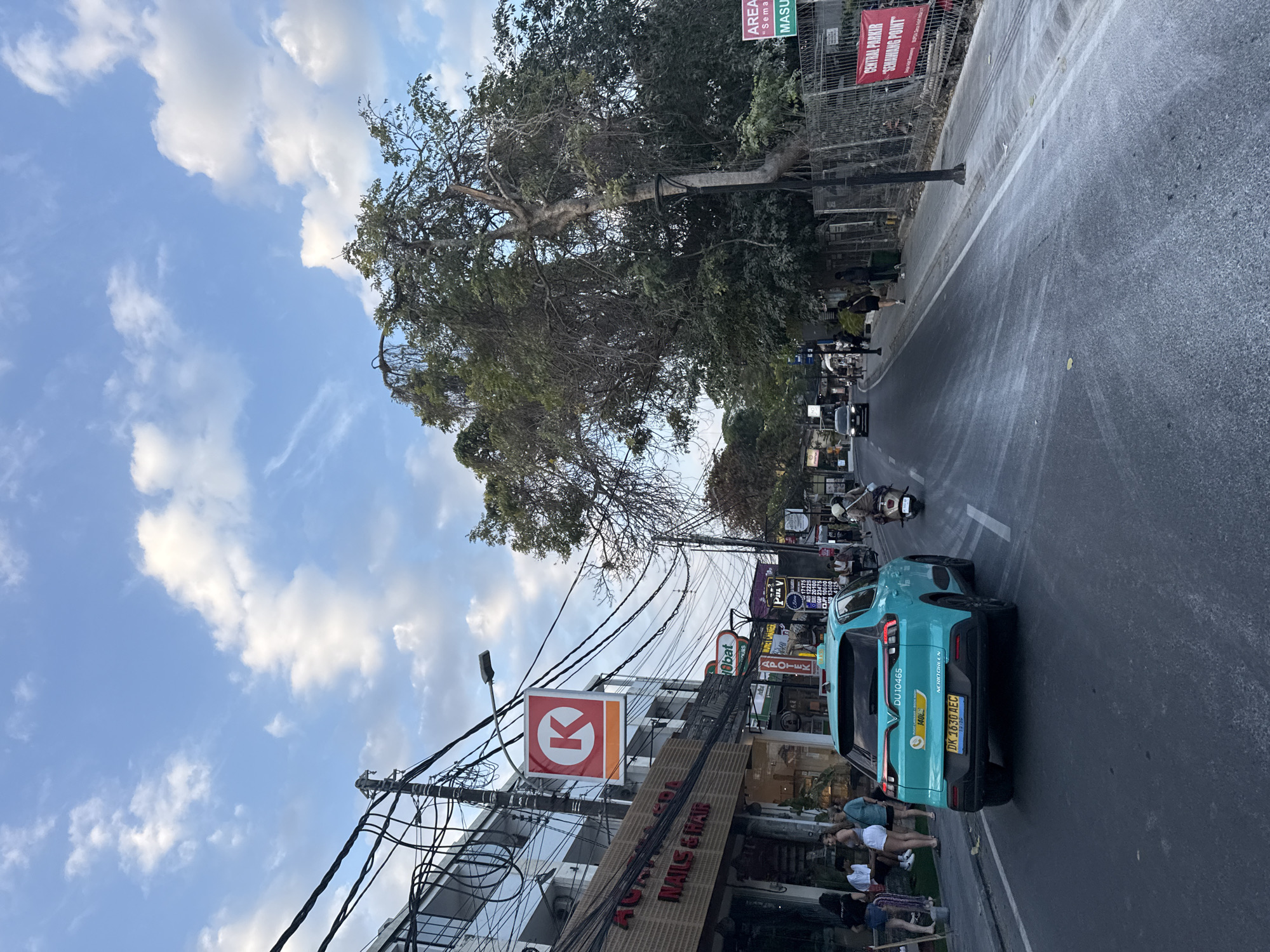 The image depicts a street scene in Denpasar, Indonesia, under a blue sky with scattered white clouds. On the left side of the street, a row of buildings houses various businesses, including "AGATHA SOUL NAILS & HAIR." A prominent red and white Circle K convenience store sign is visible. A teal-colored electric vehicle, likely a taxi, with license plate "DK 1630 AEC" is driving down the road. Several people are seen walking on the sidewalk and near the shops. On the right side of the street, a large, mature tree with dense foliage overhangs the sidewalk, and a chain-link fence borders the property. A red banner reading "CENTRAL PARKIR 'SEMAWANG POINT'" is attached to the fence. The road surface shows signs of wear and possibly some lighter-colored residue. A scooter and other vehicles are also present on the road, indicating moderate traffic. The overall atmosphere suggests a busy urban commercial area.