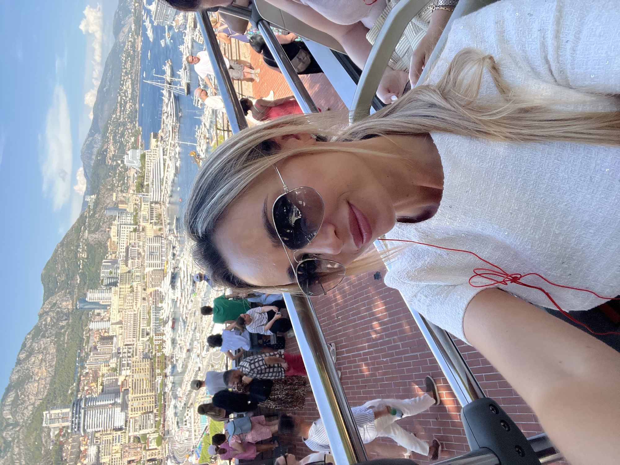 A woman with blonde hair, wearing sunglasses and a white sequined top, takes a selfie from a double-decker tour bus in Monaco. In the background, the cityscape of Monaco unfolds, featuring a dense collection of buildings cascading down a hillside towards a harbor filled with numerous yachts and boats. Mountains form a backdrop to the scene. Several tourists are visible on the upper deck of the bus and on a walkway below, observing the view. The woman's red earbuds are visible, with one earbud cord looped around her neck.
