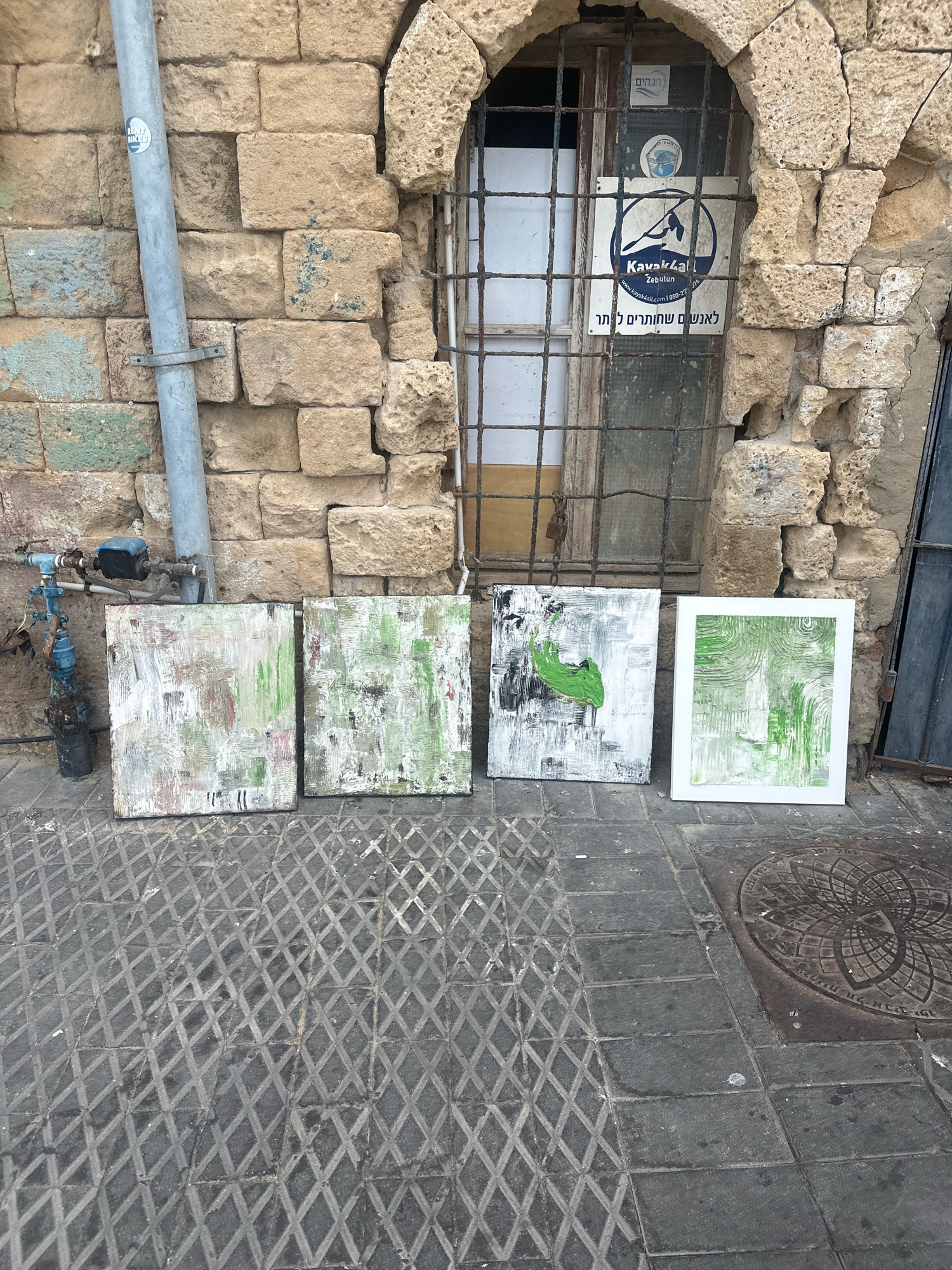 Four abstract paintings are displayed in front of a weathered stone wall in Jaffa, Tel Aviv. A metal pipe runs vertically along the left side of the wall, and a window with metal bars is visible in the center. The paintings vary in size and color, with predominantly green and white abstract patterns. The ground in front of the paintings is paved with dark grey interlocking tiles, some of which have a diamond pattern. A circular metal manhole cover with an intricate design is visible on the right side of the paved area. The overall scene suggests an outdoor art display in a historic urban setting.