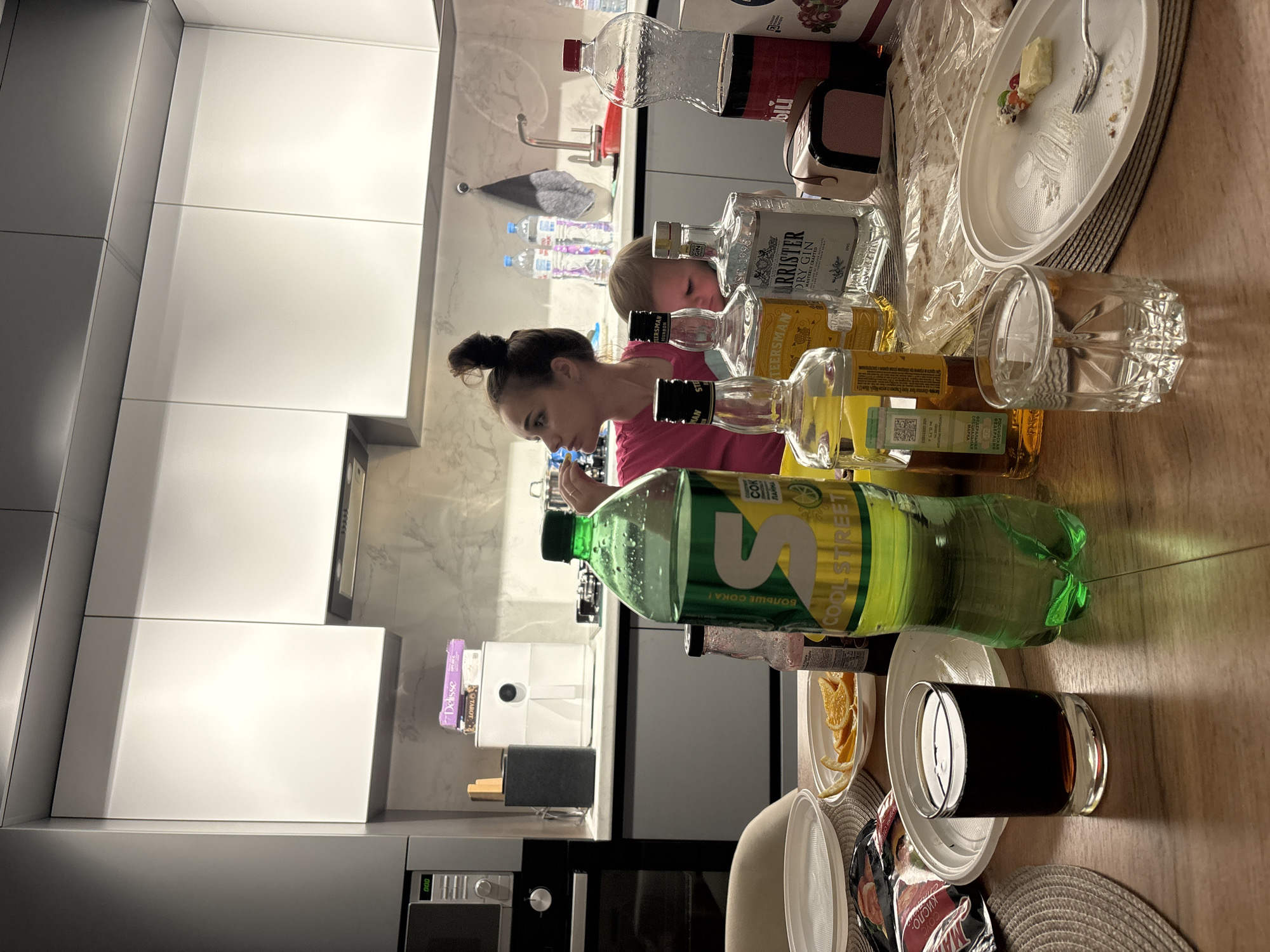 The image depicts a kitchen scene where a woman and a young child are present. The woman is seated and appears to be drinking from a green bottle of "Cool Street" soda. The child is positioned behind bottles of alcohol, observing the woman. The table is set with plates, food items such as orange slices and what appears to be a portion of cake, and several bottles of liquor including "Barrister Dry Gin" and "Steersman". In the background, kitchen cabinets are visible, along with appliances such as a microwave and an air fryer. The overall setting suggests a casual gathering. The location is specified as Шебунино, Russia.