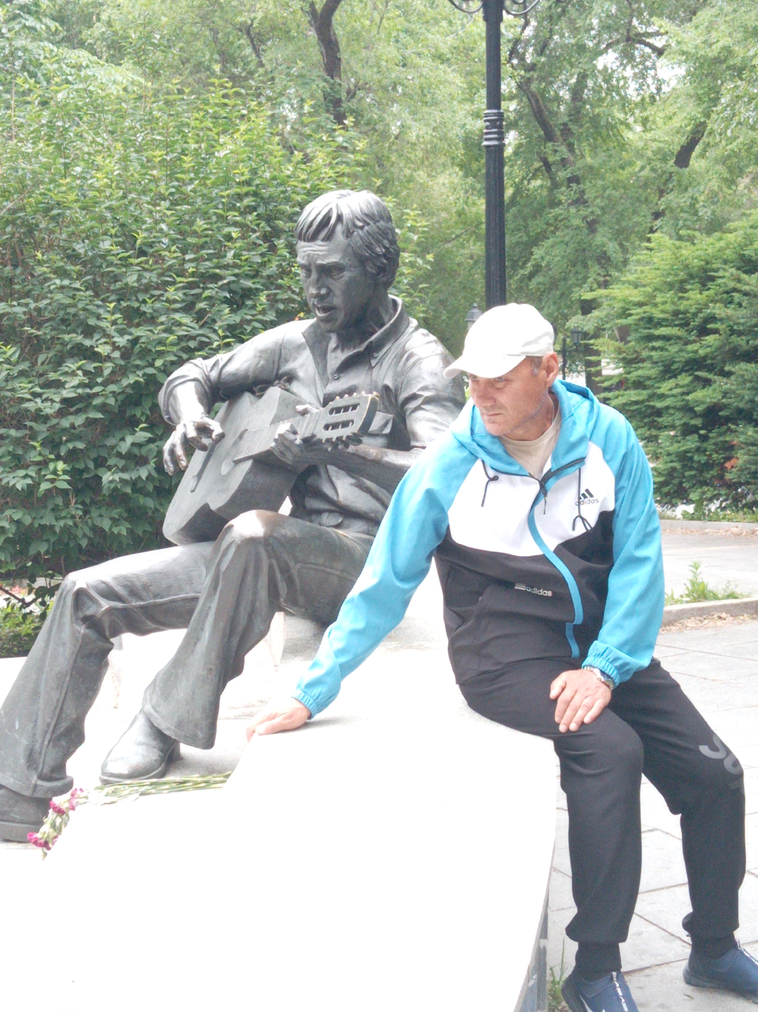 A statue of a man playing a guitar sits on a raised concrete platform in a park-like setting. The statue is made of bronze and is depicted in a seated position, holding a guitar. A living man, wearing a white baseball cap, a blue and white Adidas jacket, and black track pants, sits to the right of the statue, also on the platform. He is looking to his left, towards the statue. In front of the statue's feet, a small bouquet of pink flowers lies on the platform. The background consists of dense green foliage, trees, and a lamppost. The overall scene suggests a memorial or a place of cultural significance within a city park. The location is identified as Vladivostok, Russia.