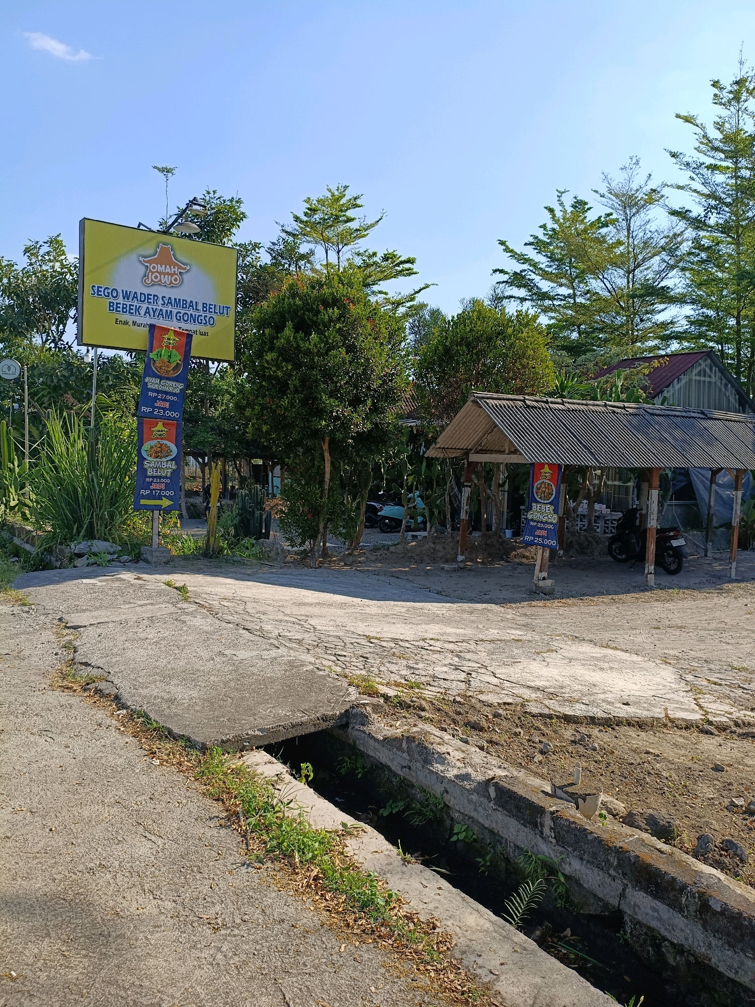The image shows a road leading towards a simple outdoor eatery in Maguwoharjo, Indonesia. A large yellow billboard for "Oma Jowo" restaurant is prominently displayed, advertising dishes like "Sego Wader Sambal Belut" and "Bebek Ayam Gongso". Next to it, a vertical sign lists prices for "Ayam Goreng Sukiyaki" and "Sambal Belut". A covered seating area with a corrugated metal roof is visible to the right, with motorcycles parked underneath. The environment is rural, with lush green trees and vegetation surrounding the establishment. A concrete drainage ditch runs alongside the road in the foreground. The sky is clear and blue.