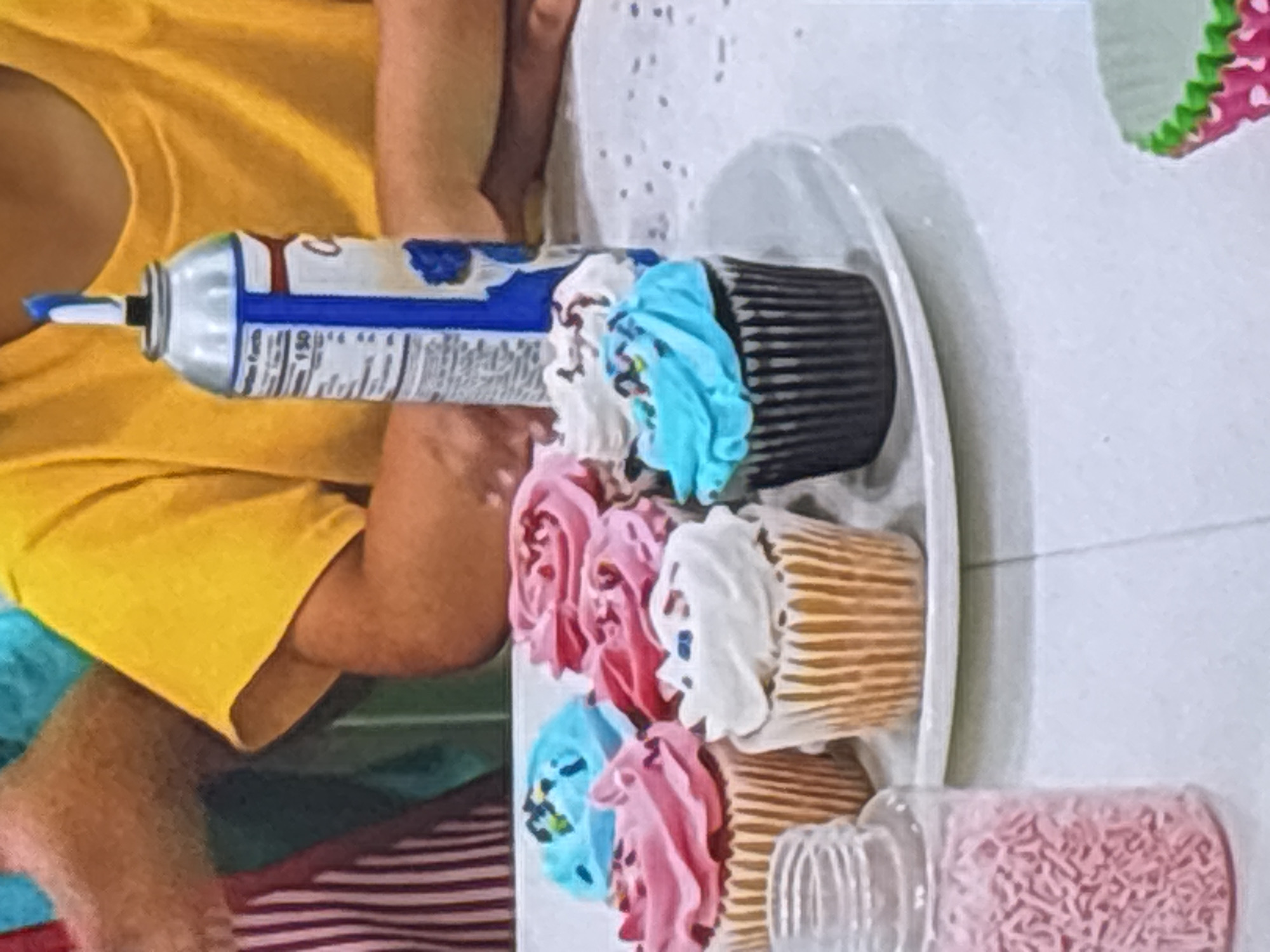 A close-up, eye-level shot shows a collection of cupcakes on a white plate, with a can of whipped cream and a jar of pink sprinkles in the foreground. The cupcakes are decorated with colorful frosting in pink, blue, and white, and some have sprinkles. Two people are partially visible in the background, wearing a striped apron and a yellow shirt, suggesting a casual gathering or party. The overall scene appears to be indoors, possibly in a home setting, on a table with a white surface. There is no information to suggest the location is in Dubai, United Arab Emirates.