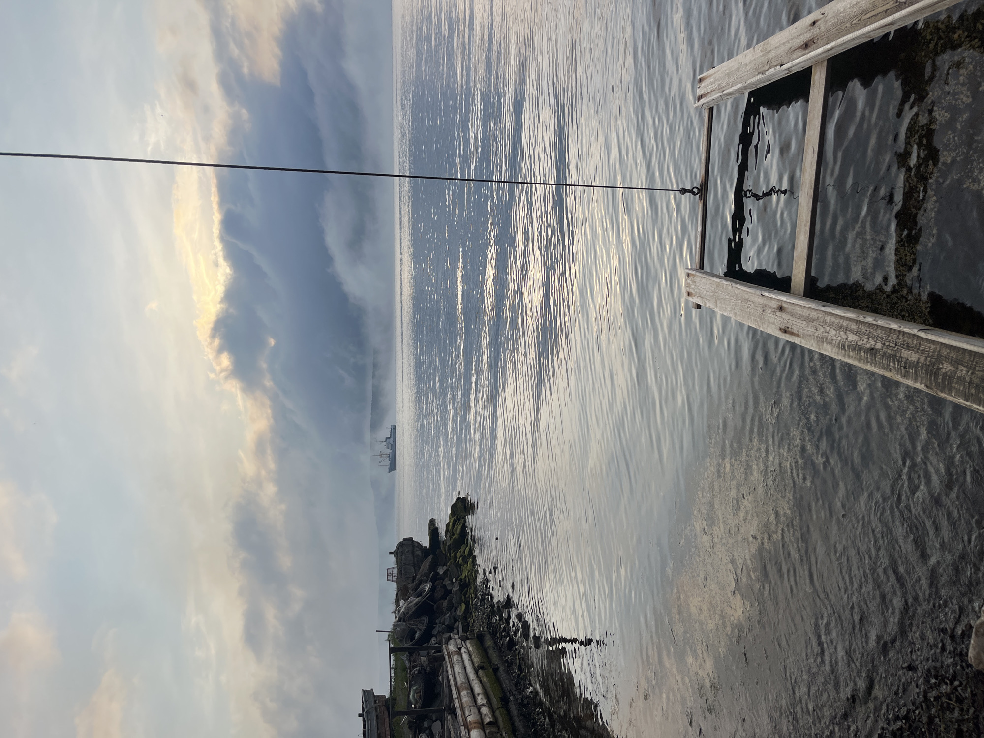 The image depicts a tranquil waterscape at what appears to be dawn or dusk, with a foggy atmosphere obscuring the horizon. In the foreground, a wooden ramp or boat launch extends into the calm water, with its lower section submerged. To the left, the shore is lined with rocks and a rustic wooden structure, possibly a pier or retaining wall, with debris such as tires and fishing gear present. In the distance, a large ship is visible on the water, partially shrouded by the fog. The sky above is a blend of soft grays and muted yellows, indicating low light conditions. The scene suggests a quiet, possibly remote, coastal location. The mention of Sovetskaya Gavan', Russia, places this scene in a port city in the Russian Far East, known for its maritime activities and scenic coastal views.