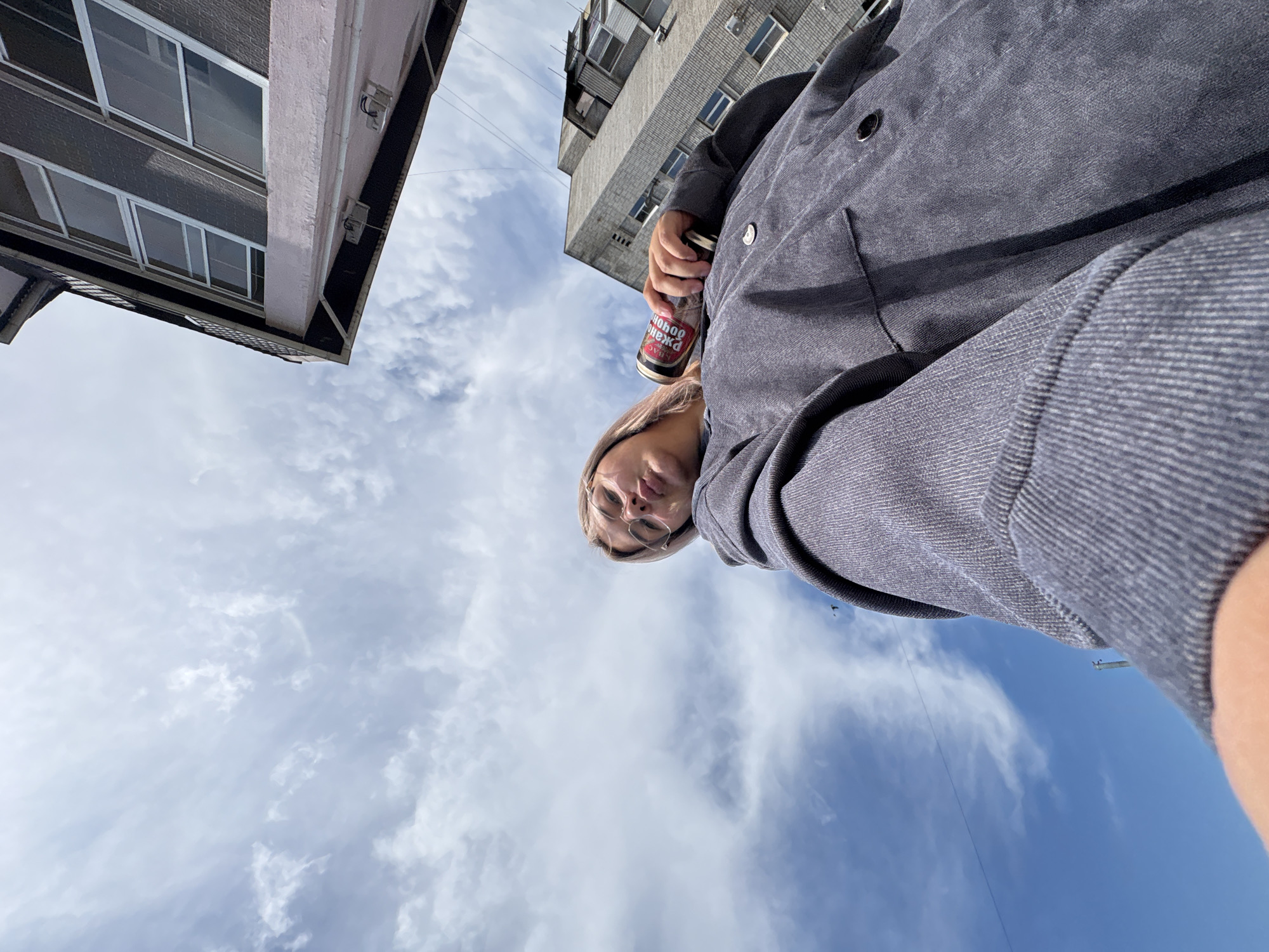 The image shows a low-angle, point-of-view shot of a woman holding a beverage can while looking up at the sky. She is wearing glasses and a dark grey jacket. In the background, there are two buildings and a cloudy blue sky. The woman's head and shoulders are visible in the foreground, with the can held in her right hand. The architecture of the buildings suggests an urban environment. The scene appears to be outdoors on a sunny day with scattered clouds.