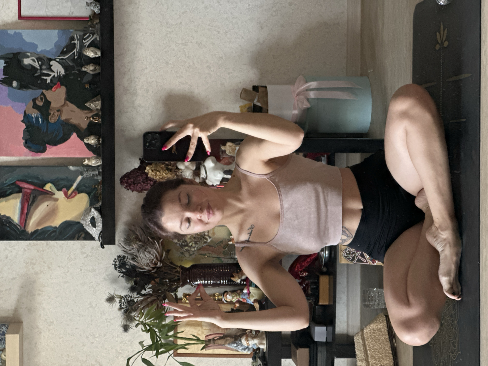 A young woman is practicing yoga in a room, likely her home, with a black yoga mat on the floor. She is sitting cross-legged, wearing a light pink tank top and black shorts. Her eyes are closed, and her hands are held in a mudra gesture. The background is cluttered with various decorative items, including paintings, figurines, and plants, suggesting a bohemian or eclectic style. The lighting appears to be natural, coming from the right side of the frame. The overall scene conveys a sense of peace and personal practice. The image does not provide any specific information about the city of Vladimir, Russia, or its locations.