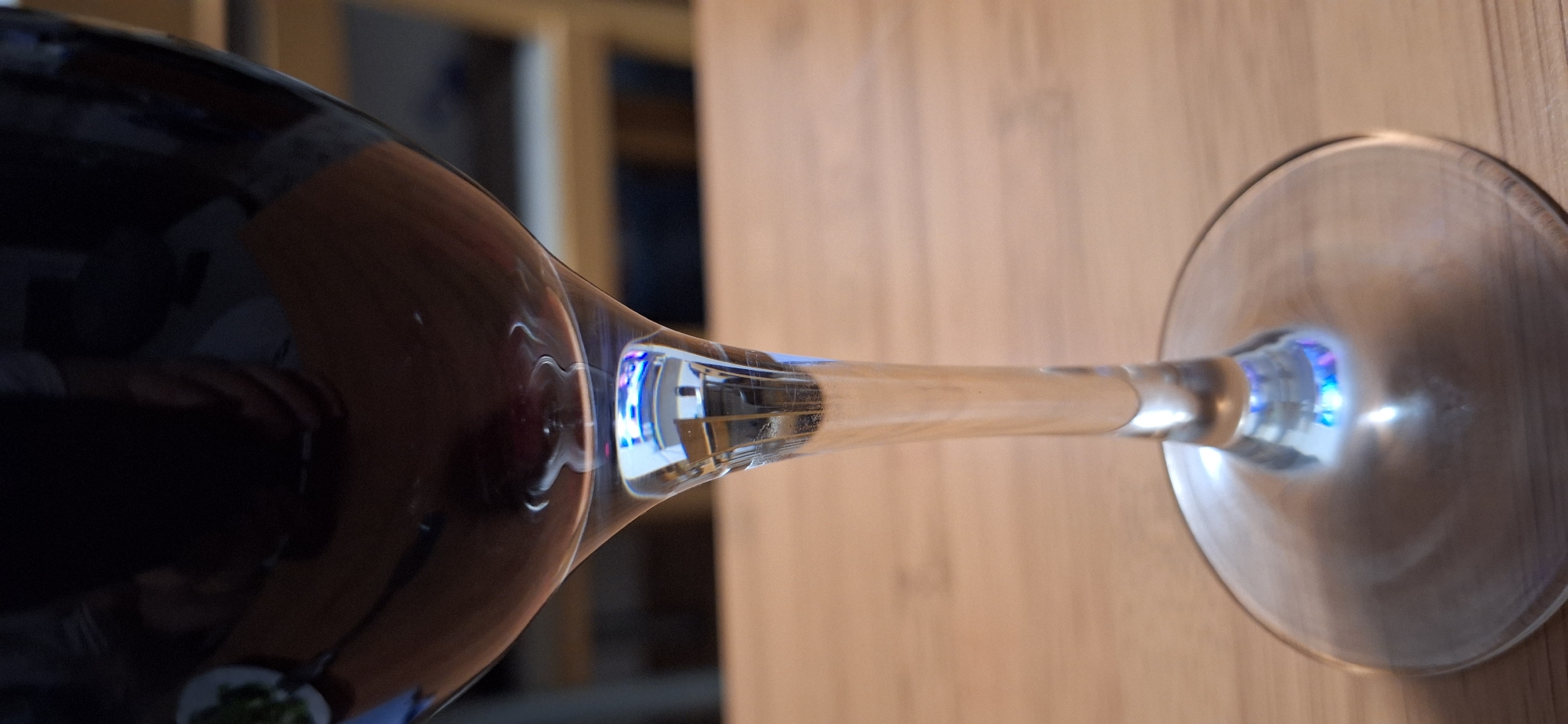 A wine glass containing red wine sits horizontally on a wooden surface. The base of the glass is to the right, and the bowl is to the left. Light appears to be refracting or reflecting within the stem and base of the glass, creating a blue hue. The background is blurry, showing what appears to be a wooden frame or furniture. The location cannot be determined from the image.