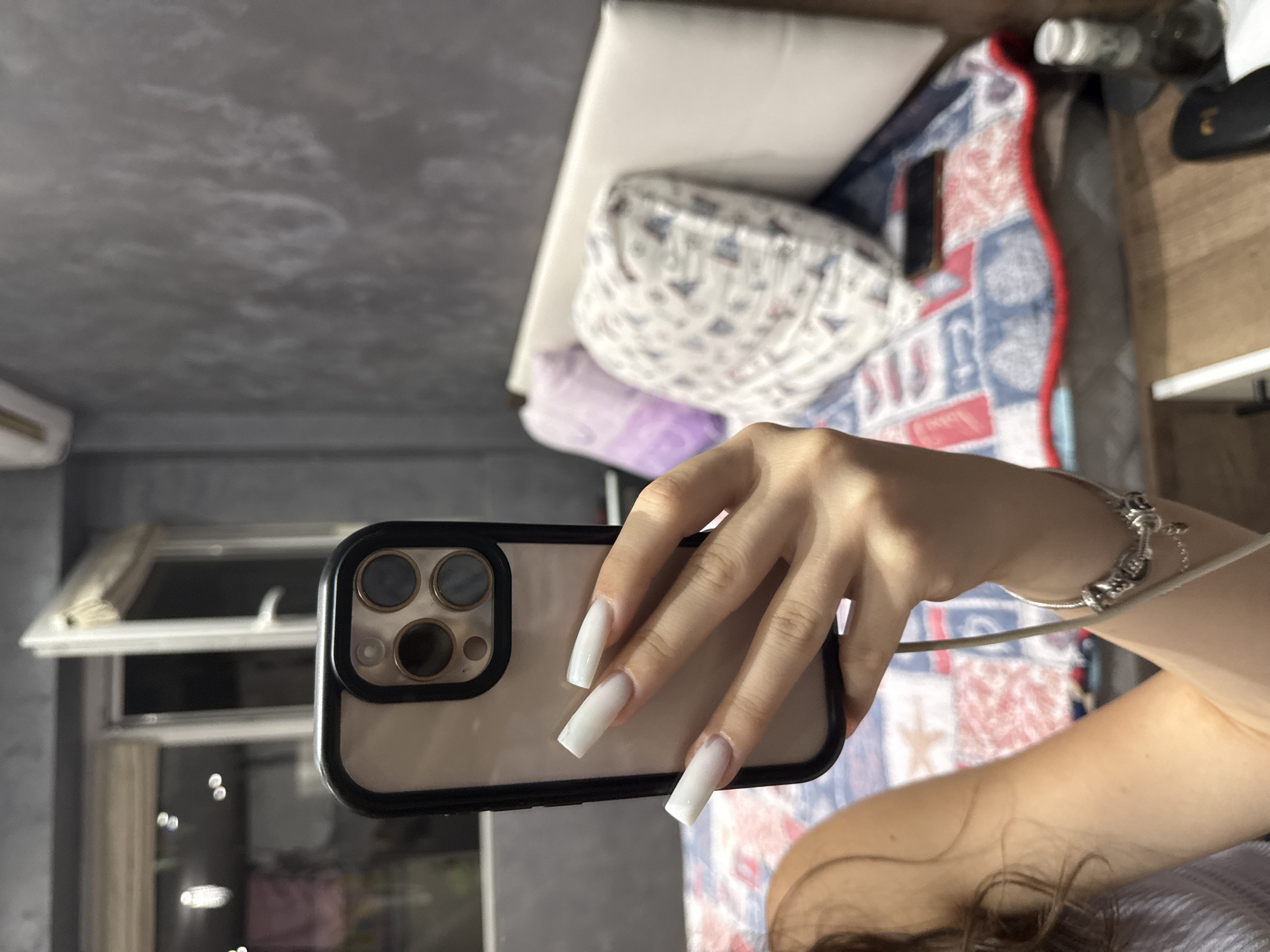 The image shows a person holding a smartphone in their right hand, taking a selfie. The person has long, square-shaped white acrylic nails. They are wearing a bracelet on their left wrist. The background appears to be a bedroom, with a bed, pillows, and a patterned duvet. A window is visible in the background, and there is a textured grey wall. A small bottle is also visible on a surface in the foreground. The image does not contain any information that would allow for identification of the location as Sofia, Bulgaria.