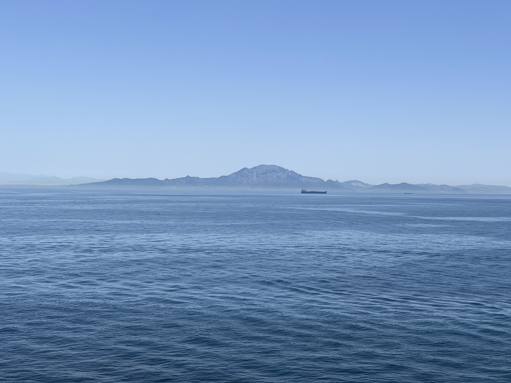 The image depicts a panoramic view of the sea with a distant mountain range. The sea is calm, with gentle ripples on its surface, reflecting the clear blue sky. In the middle distance, a large cargo ship is visible, sailing on the water. The mountain range in the background appears hazy due to atmospheric conditions, suggesting a significant distance from the viewer. The overall scene conveys a sense of openness and tranquility, characteristic of a seascape. The distinctive shape of the mountain suggests it may be the Rock of Gibraltar, a prominent landmark in Gibraltar.