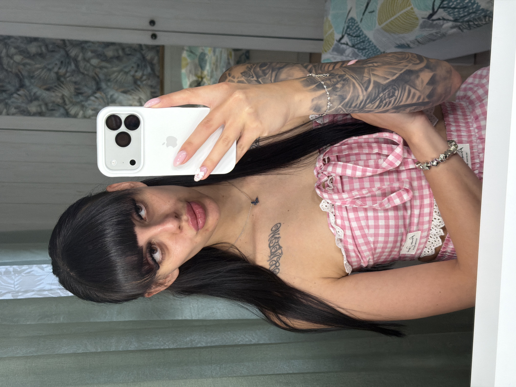 A young woman with long black hair and bangs is taking a mirror selfie in what appears to be a bedroom. She is wearing a pink and white gingham off-the-shoulder top with lace trim and a matching skirt. She has extensive tattoos on her arms and chest. In her right hand, she holds a white iPhone. A delicate silver necklace with a small blue charm is visible around her neck. The background shows a light green curtain on the left and a wardrobe with patterned doors on the right. The reflection in the mirror also reveals a bed with patterned bedding. The scene is indoors, likely a private residence. There is no information to suggest this location is in Plovdiv, Bulgaria.