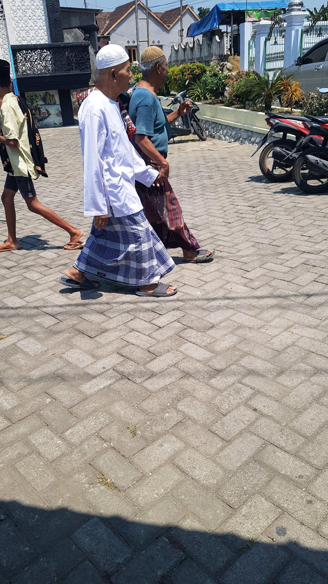 The image, captured in Kandangan, Indonesia, depicts three individuals walking on a paved walkway. In the foreground, a man wearing a white cap and a white, long-sleeved shirt with a blue and white checkered sarong walks towards the right. He is wearing dark gray slip-on sandals. To his right and slightly behind him, another man with a tan cap and a blue t-shirt is visible, also walking to the right. He is wearing a patterned sarong and dark sandals. To the left of the first man and further in the background, a young person is walking in the same direction, wearing a light yellow shirt, dark shorts, and flip-flops. In the background, a silver car is parked, and two motorcycles are also present. The walkway is made of interlocking rectangular pavers. Beyond the walkway, there are buildings with white walls and tiled roofs, and an area with lush green vegetation. The sky is clear and blue, indicating a sunny day.