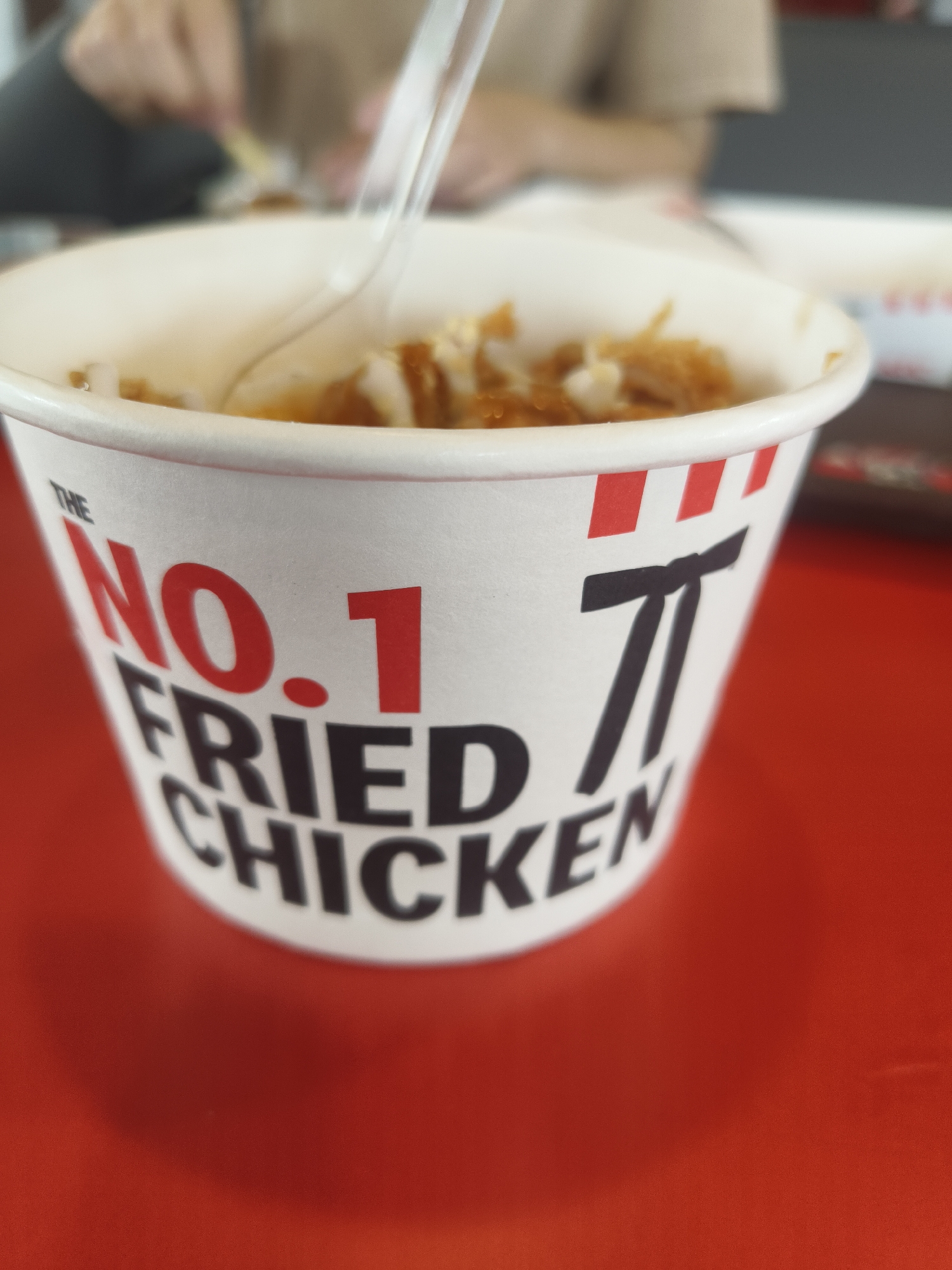 The image shows a close-up of a white paper bucket with red and white stripes around the rim. The bucket has black and red text that reads "THE NO. 1 FRIED CHICKEN". There is also a black stylized pi symbol on the side of the bucket. Inside the bucket, there is food, possibly fried chicken and rice, which appears to be partially eaten. A clear plastic fork is visible in the food. The bucket is sitting on a red surface, likely a table. In the blurred background, a person's hands are visible, holding what appears to be another utensil, suggesting they are eating. The scene is likely indoors, possibly at a fast-food restaurant in Denpasar, Indonesia, given the context of the prompt, though the specific location is not explicitly visible in the image itself. The lighting appears to be natural or overhead artificial light, casting a slight shadow.