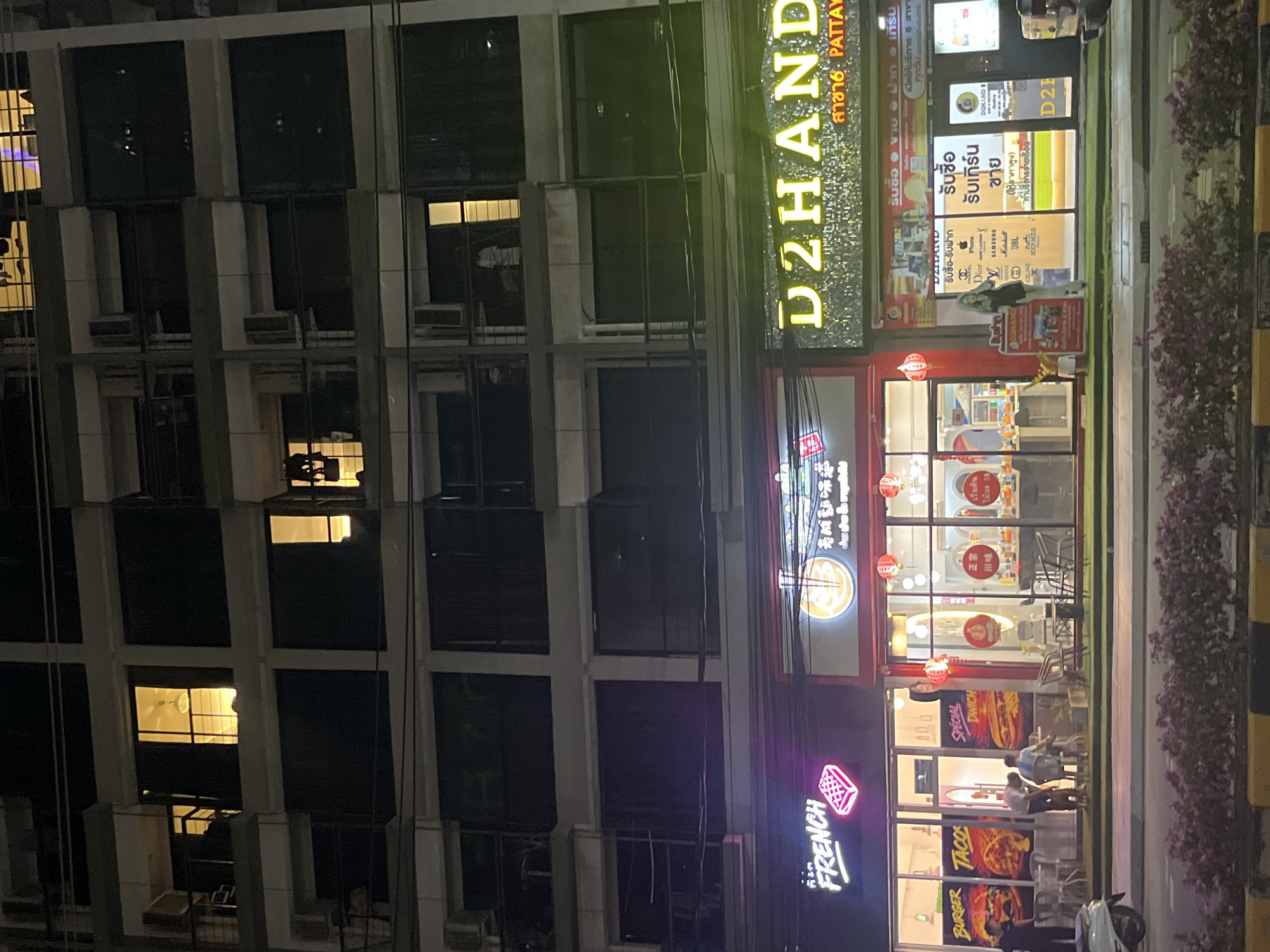 The image depicts a nighttime scene in Pattaya City, Thailand, showcasing a multi-story building alongside a commercial area. The building, constructed with concrete pillars and windowed units, is dimly lit with a few windows showing interior light. Adjacent to the building are three commercial establishments; D2HAND, a business for buying and selling items, a Chinese restaurant marked with Chinese script and red lanterns, and a fast-food restaurant named FRENCH. People are seen in front of the restaurants. The street is bordered by plants and a yellow-and-black striped curb.