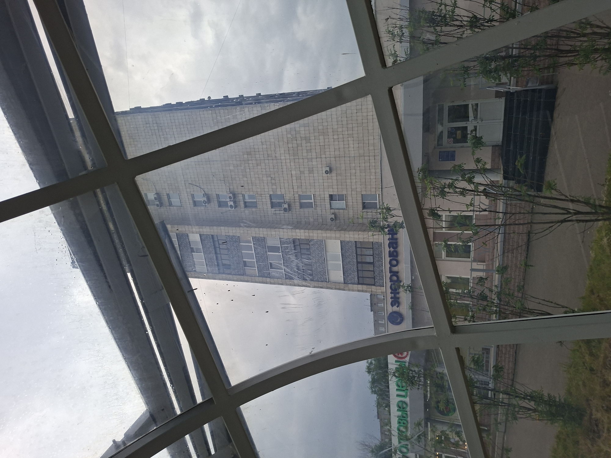 The image is a street-level, outdoor, eye-level shot of a modern building with a glass-enclosed walkway in the foreground. The building is tall, light-colored, and appears to be residential or office space. It has many windows and air conditioning units visible on its facade. Below the tall building, there is a lower, more commercial-looking structure with storefronts. Signs with Russian text are visible, including "ОТОВЫЕ ЦЕНЫ" and "Энергобанк". There are trees and plants in front of the commercial building and walkway. The sky is overcast with clouds, suggesting a cloudy or possibly rainy day. The image appears to be taken in Kazan, Russia.