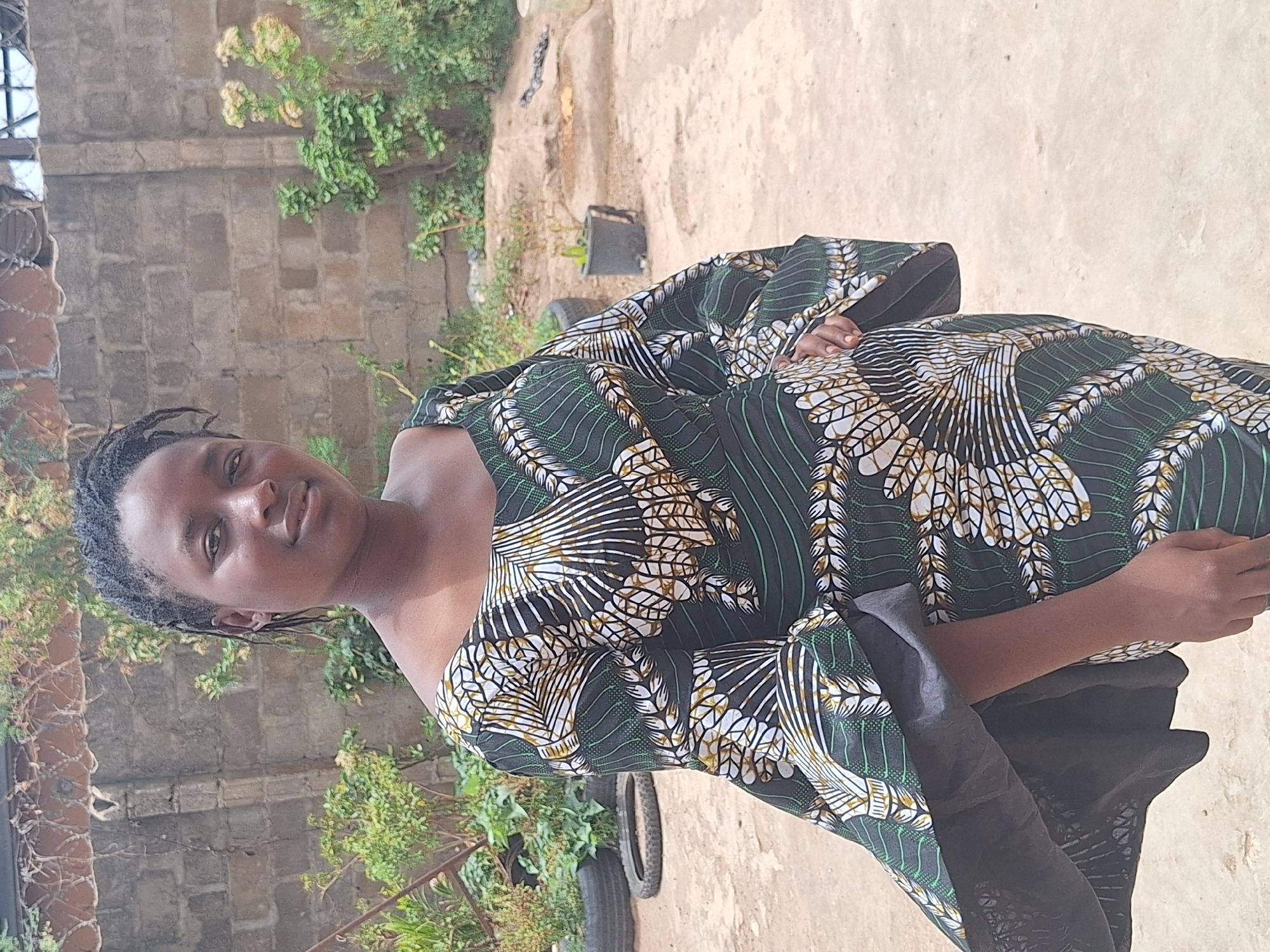 A vertical, eye-level outdoor portrait features a young woman standing and looking directly at the viewer with a calm expression and a slight smile. She has dark skin and her dark hair is styled in small braids or twists. She is wearing a vibrant, loose-fitting dress with a striking pattern, predominantly dark green, white, and golden yellow. The pattern features intricate, leaf-like or feather-like designs arranged in stripes and radiating motifs. The dress has wide, flowing sleeves, and her left hand is subtly resting near her hip.
The setting appears to be a sunny outdoor space in Sabon Tasha, Nigeria. The ground beneath her is light-colored, likely concrete or dry earth. In the mid-ground, there are discarded tires and a dark potted plant with green foliage. Behind her and to her left is a weathered, rough stone or concrete block wall. Lush green foliage and some lighter-colored plants are scattered around the background, suggesting a natural or semi-natural environment. Bright natural light illuminates the scene, indicating it is daytime with clear weather. There is no visible text in the image. The overall mood conveyed by the subject is serene and pleasant.