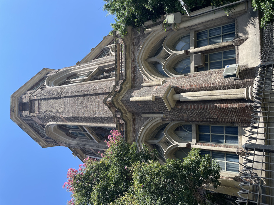 The image displays a tall, ornate building, likely a tower or a significant section of a larger edifice, captured from a low angle looking upwards against a clear blue sky. The building exhibits characteristics of Gothic Revival architecture, featuring dark red brickwork contrasted with lighter-colored stone or concrete trim and decorative elements.
The structure is multi-storied and vertical, with prominent architectural features. Several windows are visible, some designed with pointed Gothic arches and intricate tracery patterns, including trefoil shapes. Other windows are rectangular with multiple panes, many appearing to have blue-tinted or reflective glass. Decorative elements such as columns, pilasters, cornices, and buttress-like projections adorn the facade.
Modern additions are evident on the historical building, including two white air conditioning units mounted below rectangular windows on the exterior brickwork. A white floodlight fixture is also attached to an upper section of the building.
Green foliage from large trees, interspersed with clusters of pink blossoms, partially frames and obscures the lower and mid-sections of the building, adding a touch of natural color. Along the right side of the image (which would be the lower-right when viewed upright), a dark, spiky metal fence or railing is visible.
The sky is a bright, unblemished blue, suggesting clear weather and strong sunlight. The lighting creates defined shadows that emphasize the texture of the brick and the depth of the architectural details. No discernible text is visible anywhere in the image. The scene captures a blend of historical architecture with modern urban elements.