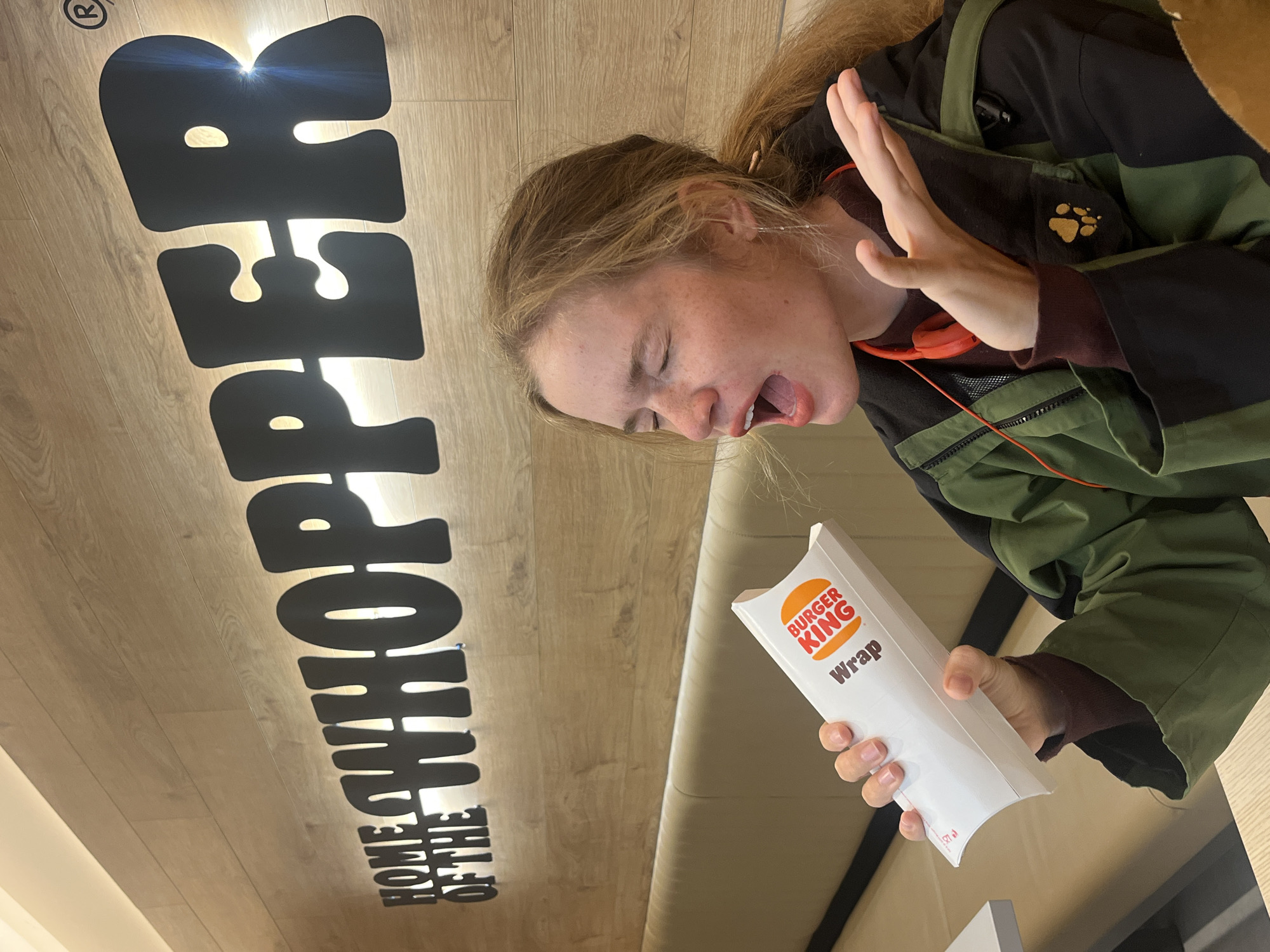A young, fair-skinned person stands in a Burger King in Budva, Montenegro. The person, with light brown hair, wears a green jacket and appears to be recoiling from a Burger King wrap held in their hand. Their mouth is open in an expression of distaste or surprise. Above the person is a sign that reads "HOME OF THE WHOPPER" in large, black lettering against a wooden surface. The lighting in the Burger King is bright and modern.