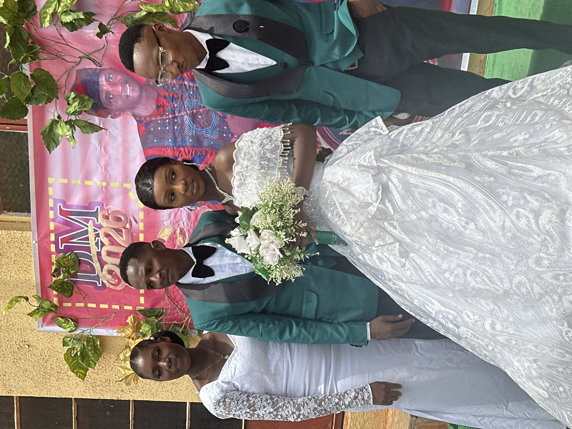 The image shows a group of four people posing in front of a backdrop. In the foreground, there are two women and two men. One of the women wears a white wedding gown with lace details, and the other woman wears a similar white dress with lace sleeves. One of the men wears a green suit with a black bow tie, while the other man wears a similar suit but with glasses. The woman in the wedding gown is holding a bouquet of white flowers. Behind them, there is a backdrop with green leaves and a poster that reads "DM 2026". The location is in the city of Unknown, Unknown.