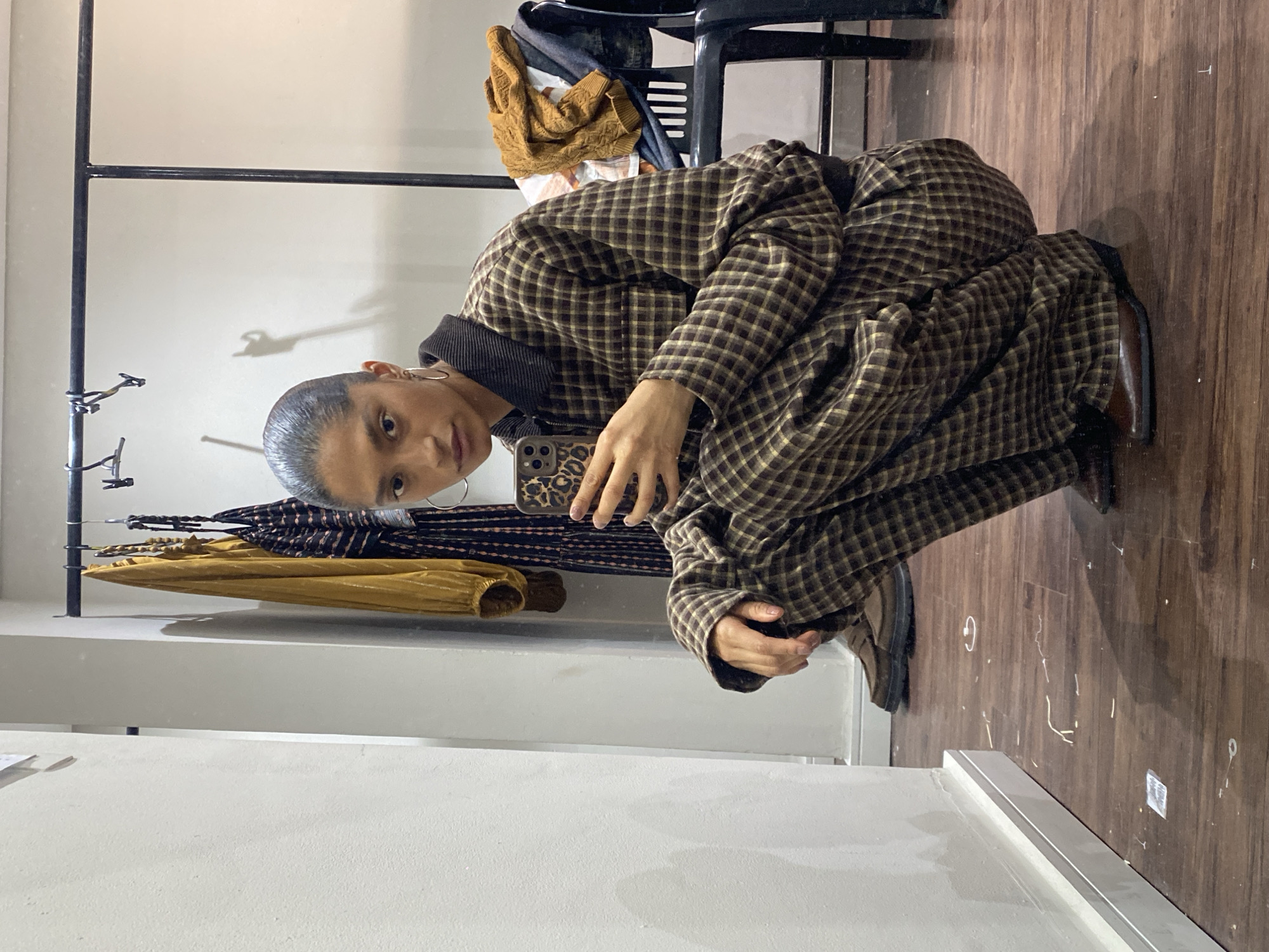 The image depicts a person crouching in what appears to be a fitting room. The individual has short, dark hair, visible hoop earrings, and is holding a smartphone with a patterned case. They are wearing a brown and yellow plaid jacket. Clothes are hanging on a rack behind them. The room has light-colored walls and dark wooden flooring. A black chair is also visible in the background. The image does not contain location information that can be used to identify the setting as being in São Paulo, Brazil.