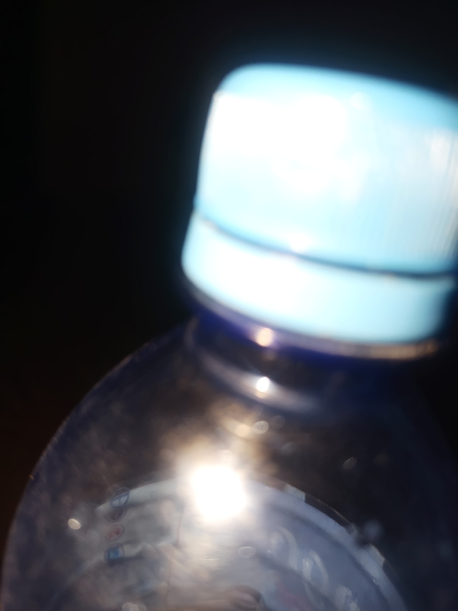 The image is a close-up, blurred, and slightly overexposed shot of the cap and upper portion of a blue plastic water bottle against a dark background. The cap is light blue and appears to be made of plastic. A bright light source is reflecting off the bottle, creating glare and obscuring details. The background is predominantly black. The scene appears to be indoors or in a location with controlled lighting, possibly a studio setting or a dimly lit room. No specific location in a city is identifiable.