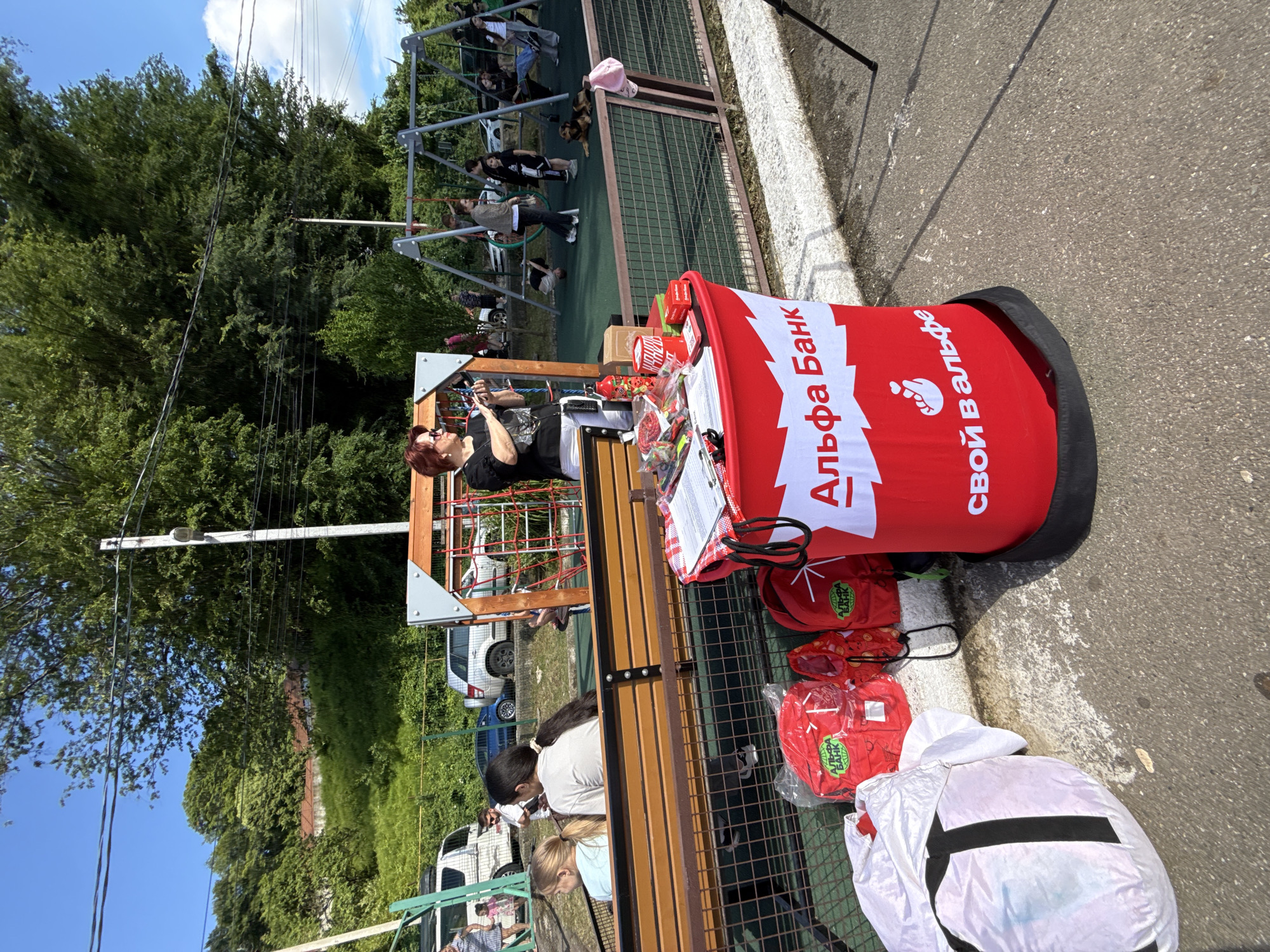 In Prohress, Russia, an outdoor event is taking place near a playground. A red and white branded table displays various items, including pamphlets and promotional materials. Several red backpacks with "Alpha Bank" branding are placed on the ground near the table. A woman with red hair, wearing sunglasses and a black t-shirt, stands behind the table, holding a camera or phone. Children are visible in the background, some playing on the swings and playground equipment. There are cars parked in the background and trees and a grassy hillside on the left side of the image. The ground is paved with asphalt and concrete.