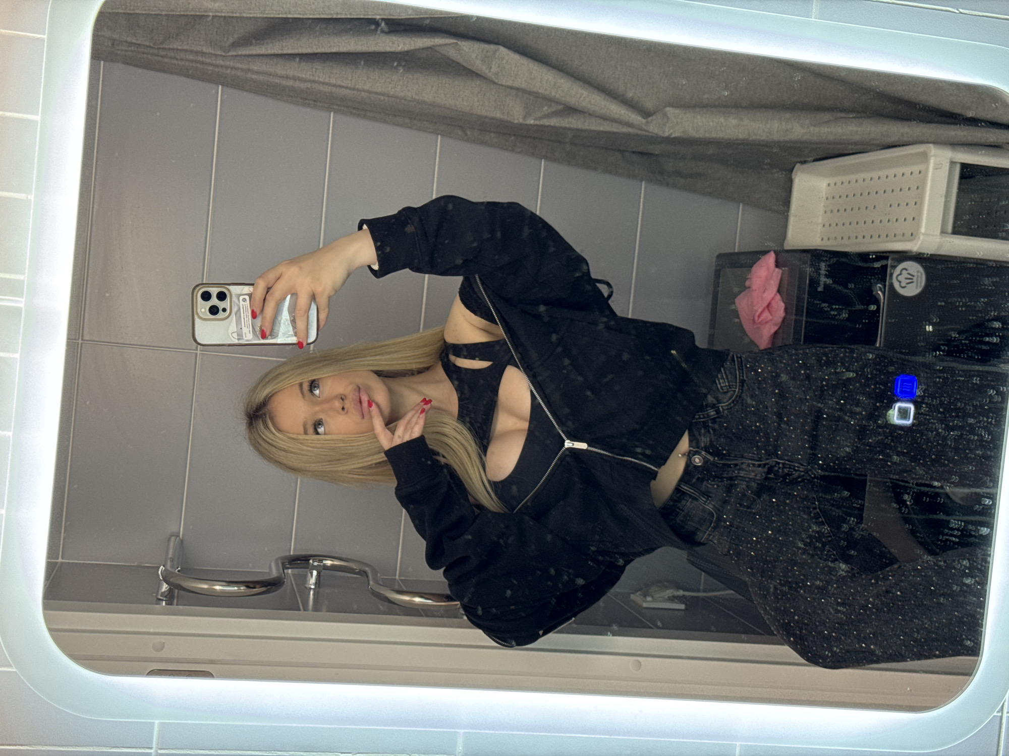 A young woman with blonde hair takes a mirror selfie in what appears to be a bathroom. She is wearing a black crop top with a zipper and cutouts, paired with dark, sparkly jeans. Her nails are painted red. She is posing with her hand to her face and looking at the camera. Behind her, a gray tiled wall and a large, illuminated mirror are visible. In the reflection, we can see some laundry baskets and a pink cloth. The scene is brightly lit, suggesting daytime or good artificial lighting. The overall mood is casual and playful. There is no visible text in the image.