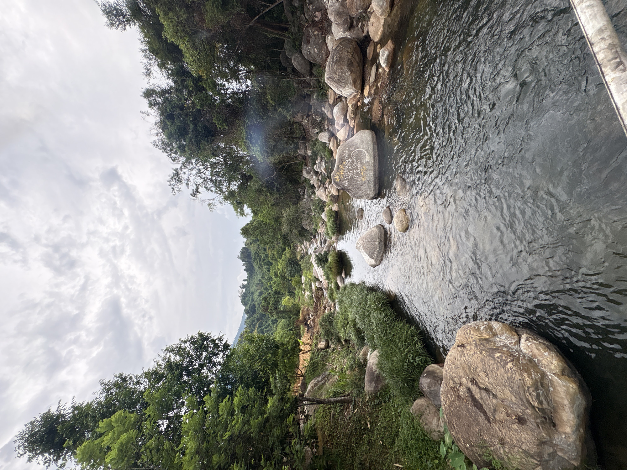 The image shows a stream in Khanh Vinh, Vietnam. Large rocks are positioned in and around the stream. Dense green vegetation surrounds the stream. A mountain slope covered in trees rises in the distance. The sky is overcast.