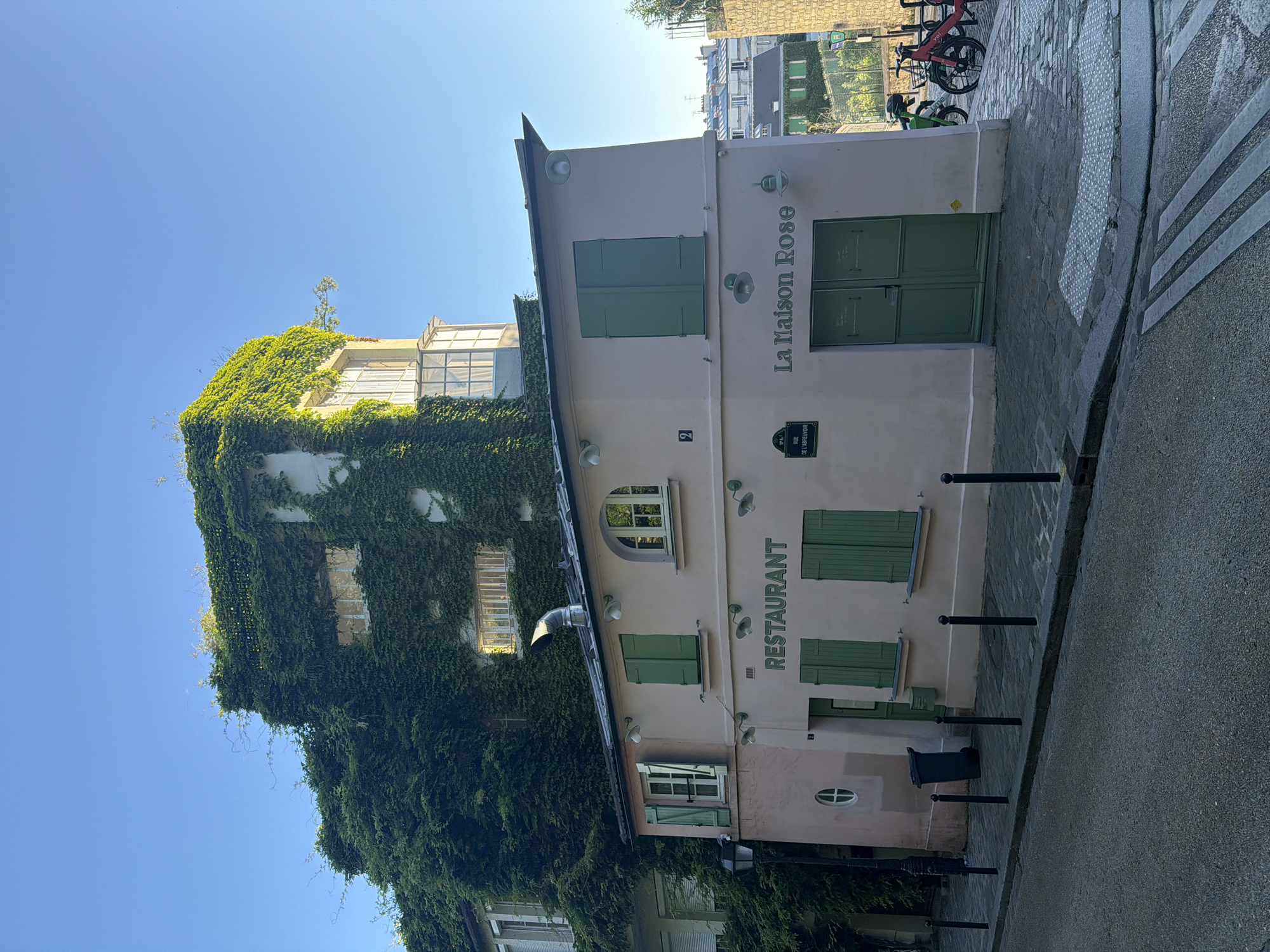 The image depicts La Maison Rose, a restaurant in the Montmartre neighborhood of Paris, France. The building is a multi-story structure with a pale pink facade. Green shutters adorn the windows. Lush green ivy covers a significant portion of the building's exterior. Signage indicates "La Maison Rose" and "Restaurant". A street sign is visible. The sky is clear and blue. The street in front of the building is made of cobblestones, with modern road markings and bollards visible. Bicycles are parked nearby. The scene appears to be captured during daylight hours.