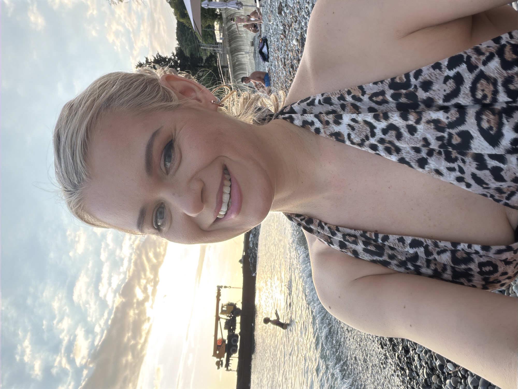 A blonde woman, wearing a leopard-print bikini top, smiles at the camera on a pebble beach in Sochi, Russia. In the background, the Black Sea is visible with a child playing in the water. A construction crane is also seen on the horizon. Several people are relaxing on the beach behind her, with umbrellas and lounge chairs set up. The sky is filled with clouds, and the sun is setting, casting a warm glow over the scene.