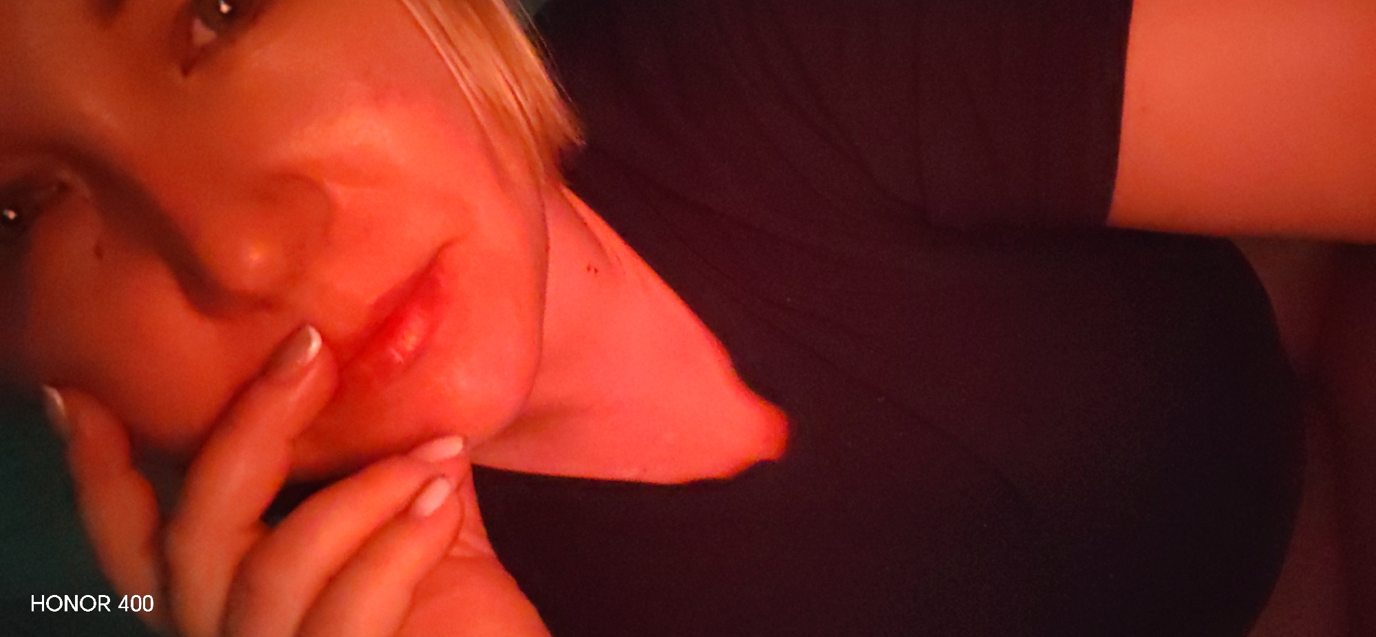 The image presents a close-up, diagonally oriented portrait of a single individual, identified as a female, illuminated predominantly by a red light source. The subject's face occupies the left and central portions of the frame, showing partial views of both eyes, a prominent nose, and full lips. Light-colored hair is visible on the upper-left. Her right hand, featuring light-colored nail polish, rests against her chin and lower cheek. She is wearing a dark-colored garment, visible on her upper chest and shoulder area. The strong red illumination casts a reddish-orange hue across her skin. The background is dark and indistinct. The text "HONOR 400" is visible in the bottom-left corner. The specific location within the city of Noviy, Russia, cannot be ascertained from the visual evidence.