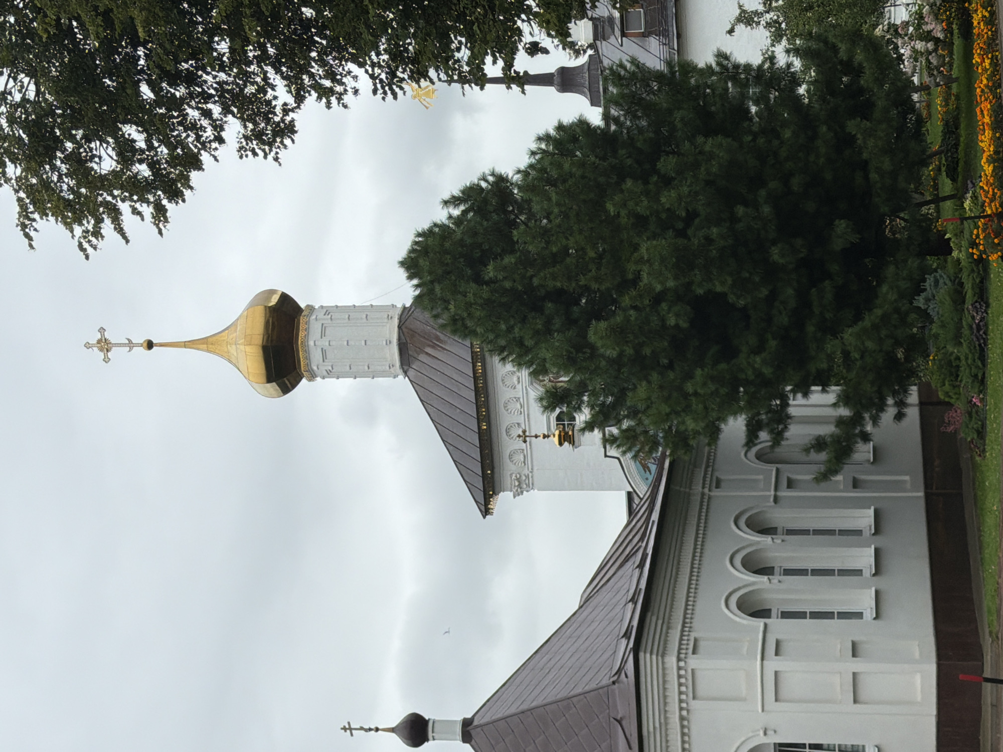The image depicts an exterior view of a white building with arched windows and decorative molding, likely a church or monastery, in Rusanovo, Russia. A large evergreen tree dominates the foreground, obscuring a portion of the building. In the background, a white tower with a golden dome and cross is visible, along with a darker, multi-gabled roof. The sky is overcast, with scattered clouds. The ground in front of the building is covered with grass and flower beds containing orange and yellow flowers. The architectural style suggests a religious or historical significance. There are no people visible in the image.
